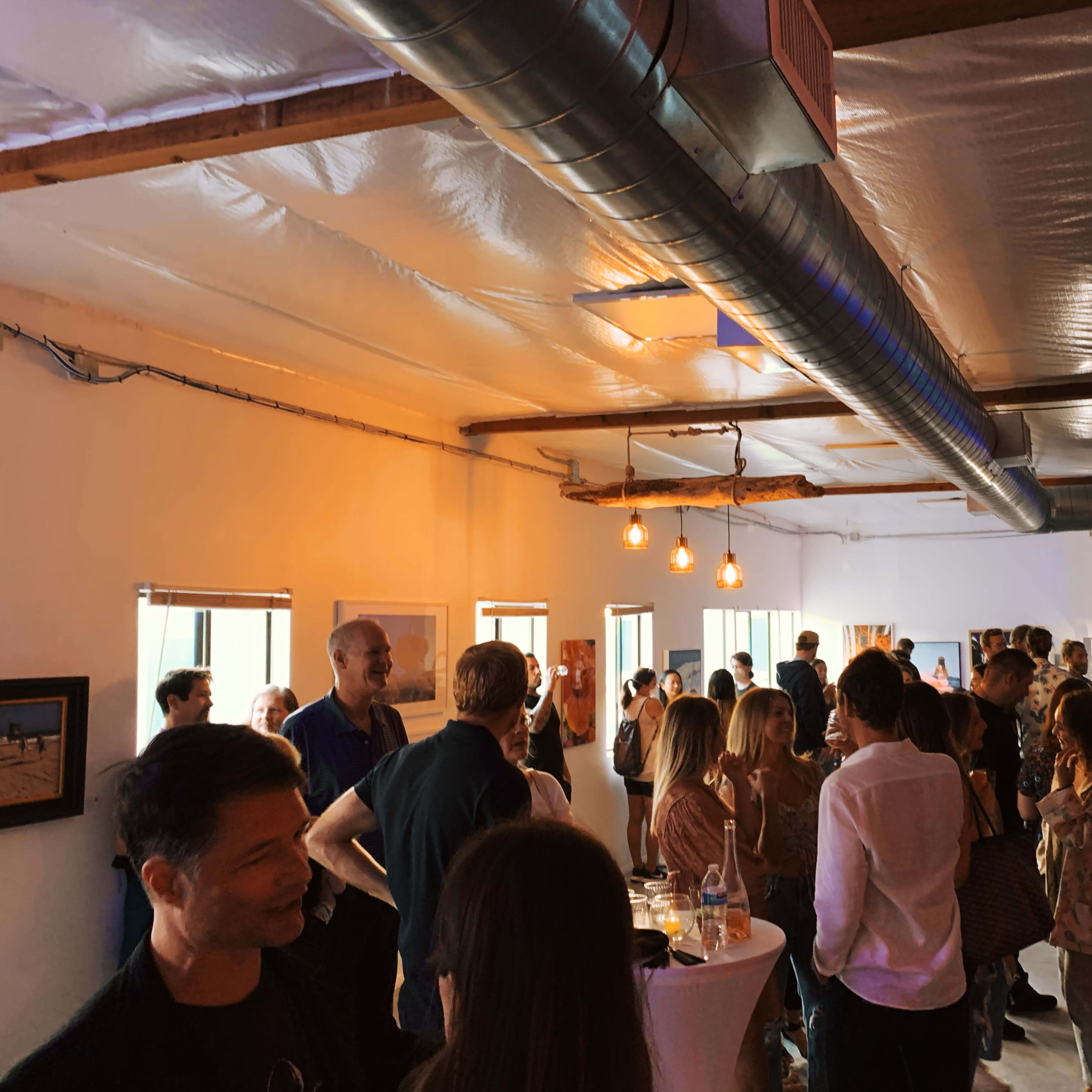 A crowded indoor space featuring people socializing and engaging at a gathering, with exposed beams and hanging lights.