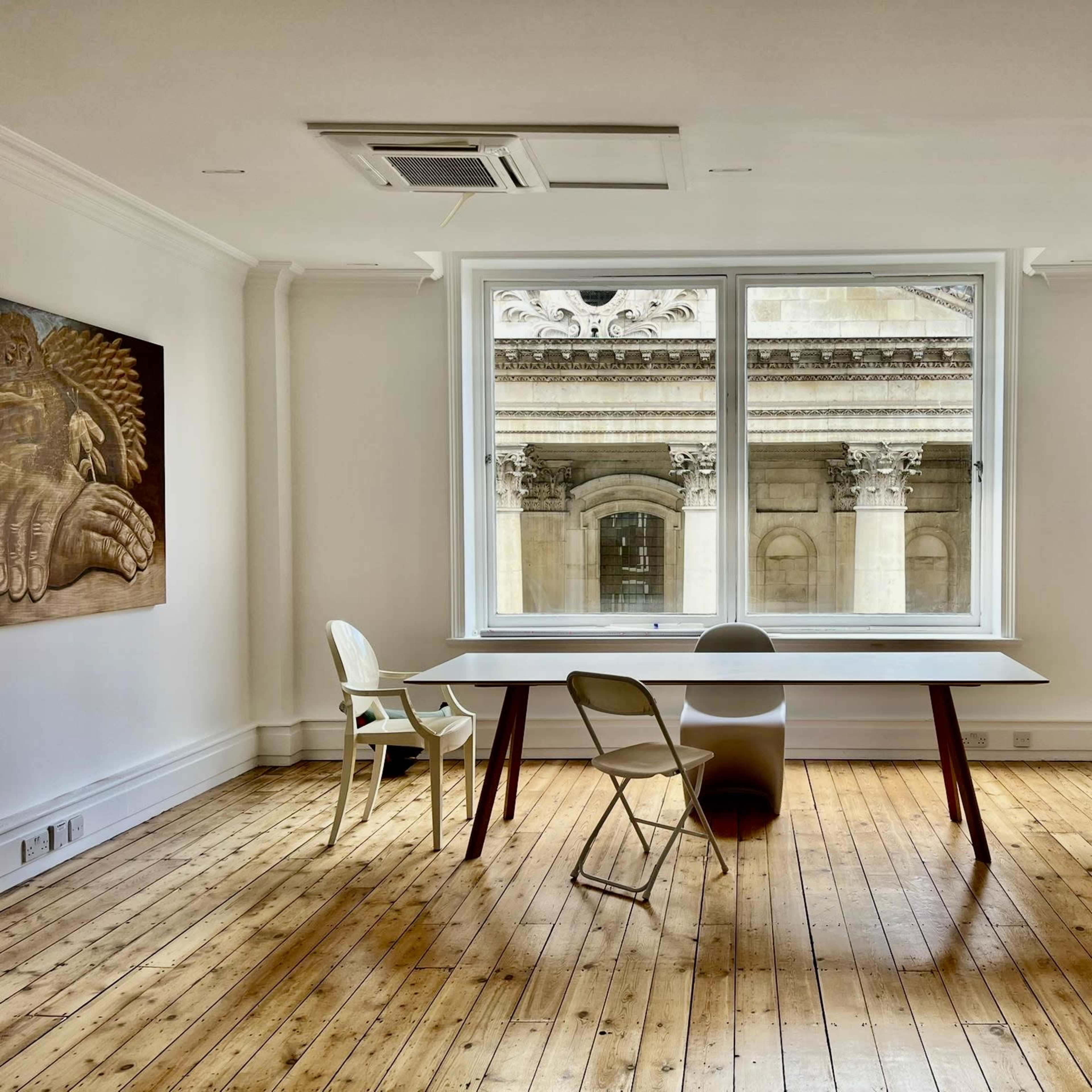 A minimalist room features a large window overlooking a historic facade, with a table and chairs arranged on wooden flooring.