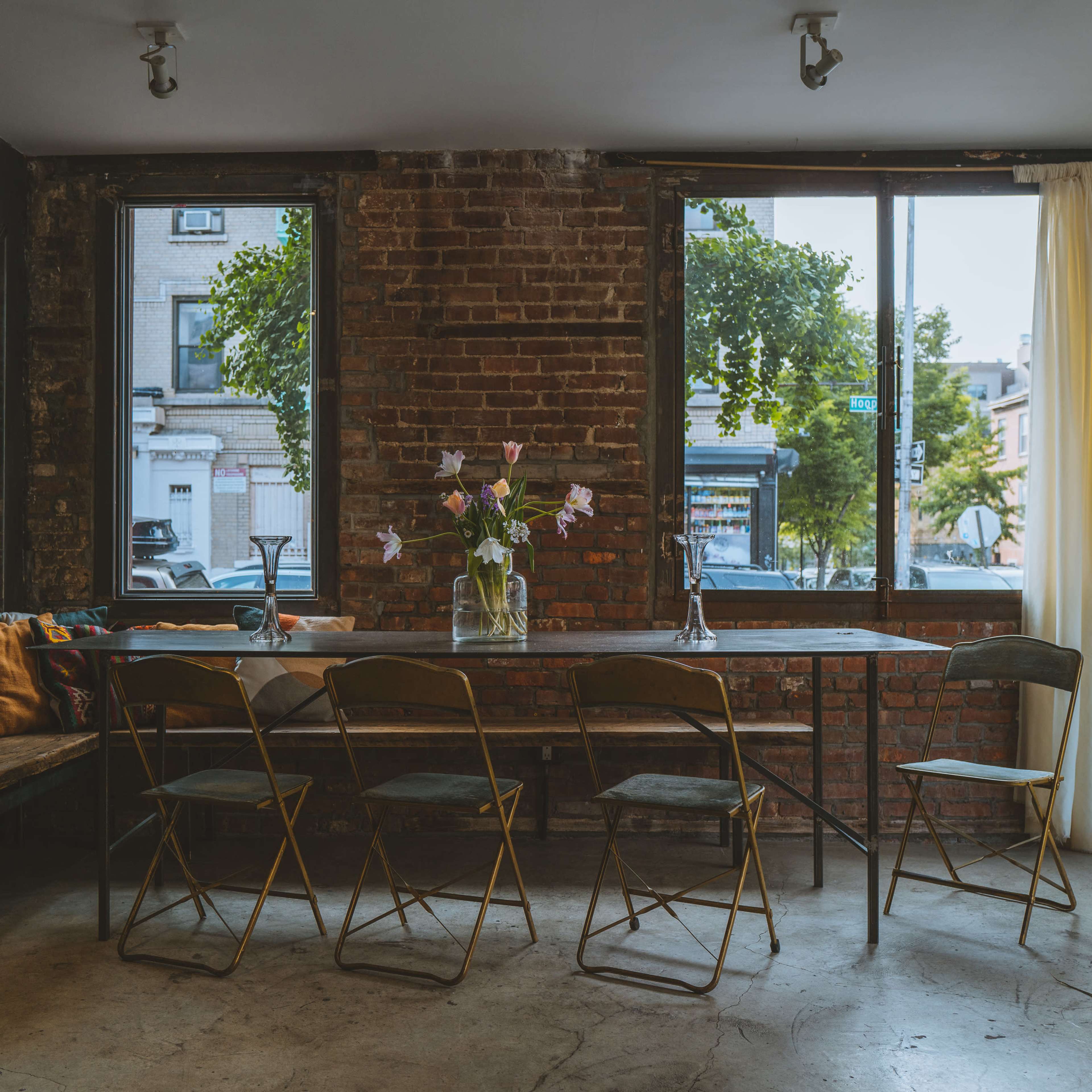 A long table with gold chairs sits in a room featuring exposed brick walls and large windows that let in natural light, complemented by a vase of flowers at the center.
