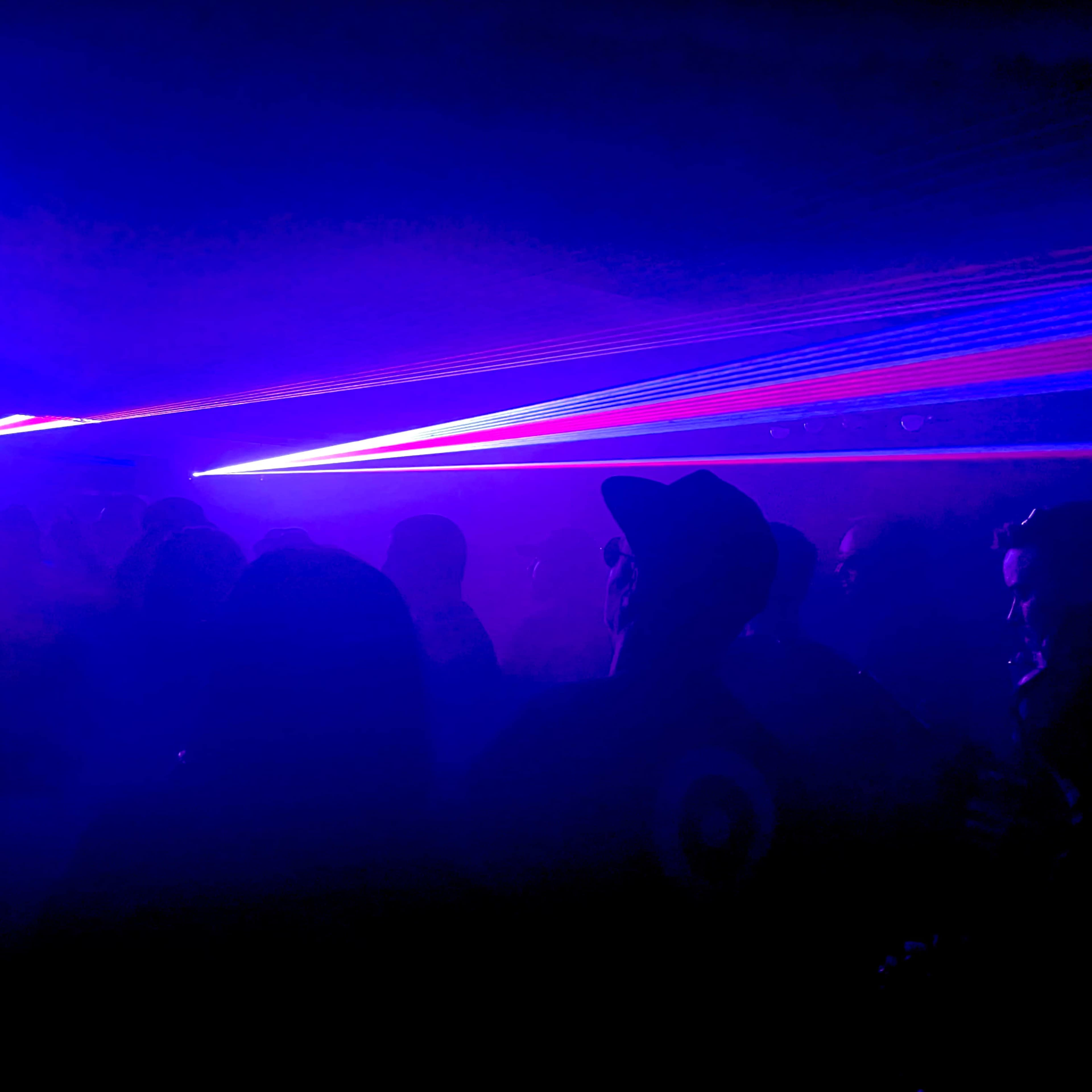 A crowd of people is silhouetted against colorful laser lights in a dimly lit space.