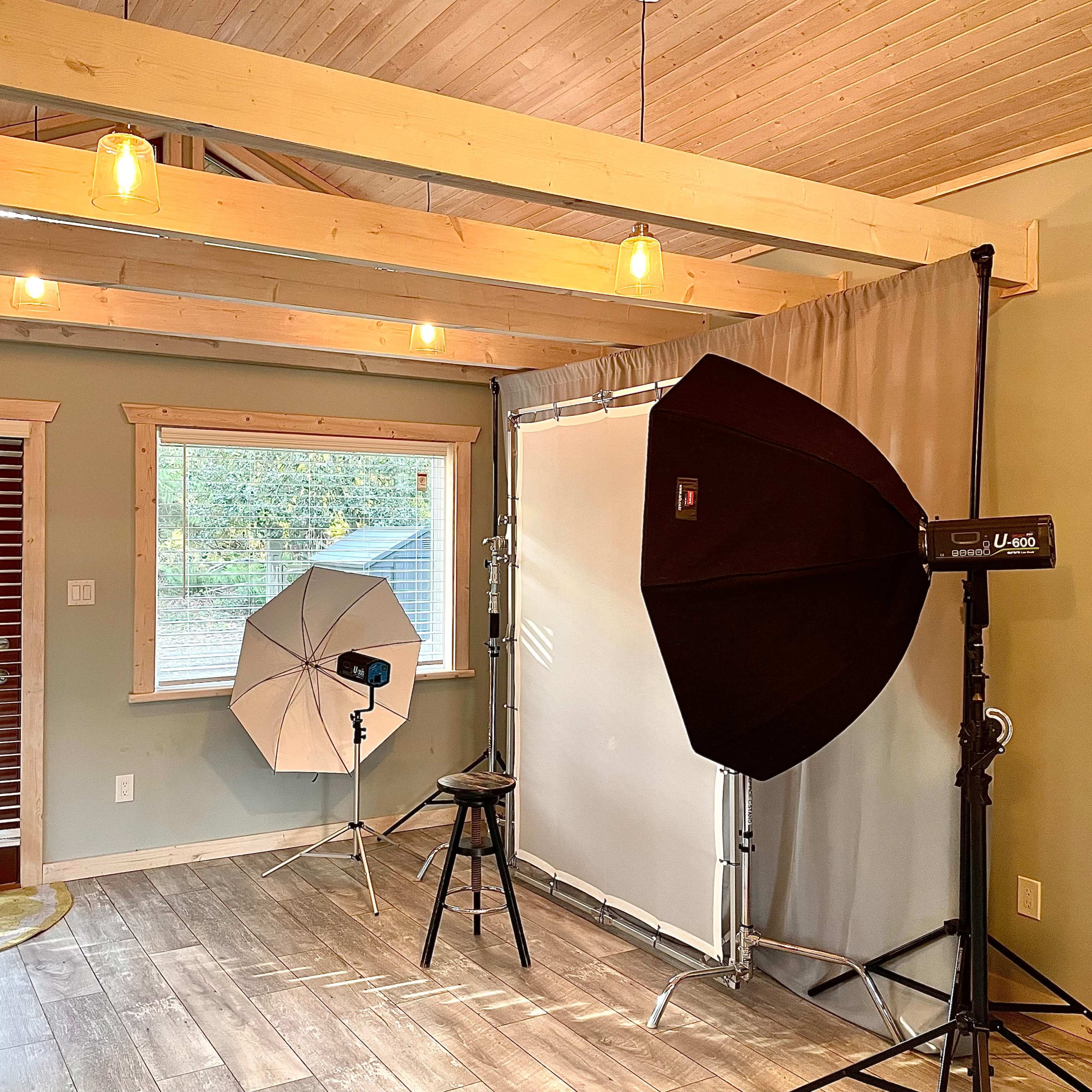 A photography studio setup featuring a backdrop, lighting equipment, and a stool in a room with wooden beams and large windows.