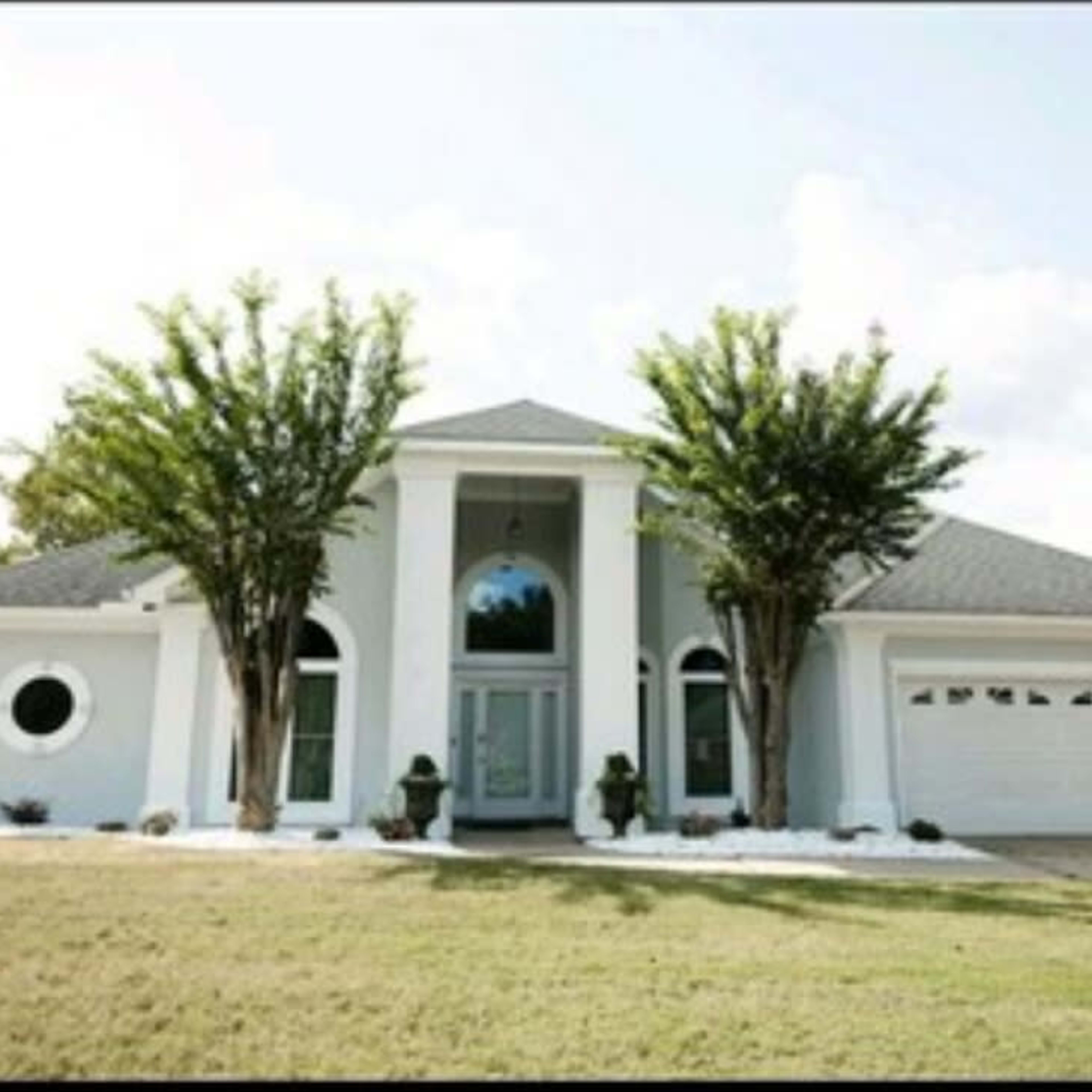A modern, single-story house with a symmetrical facade, large front windows, and two trees flanking the entrance.