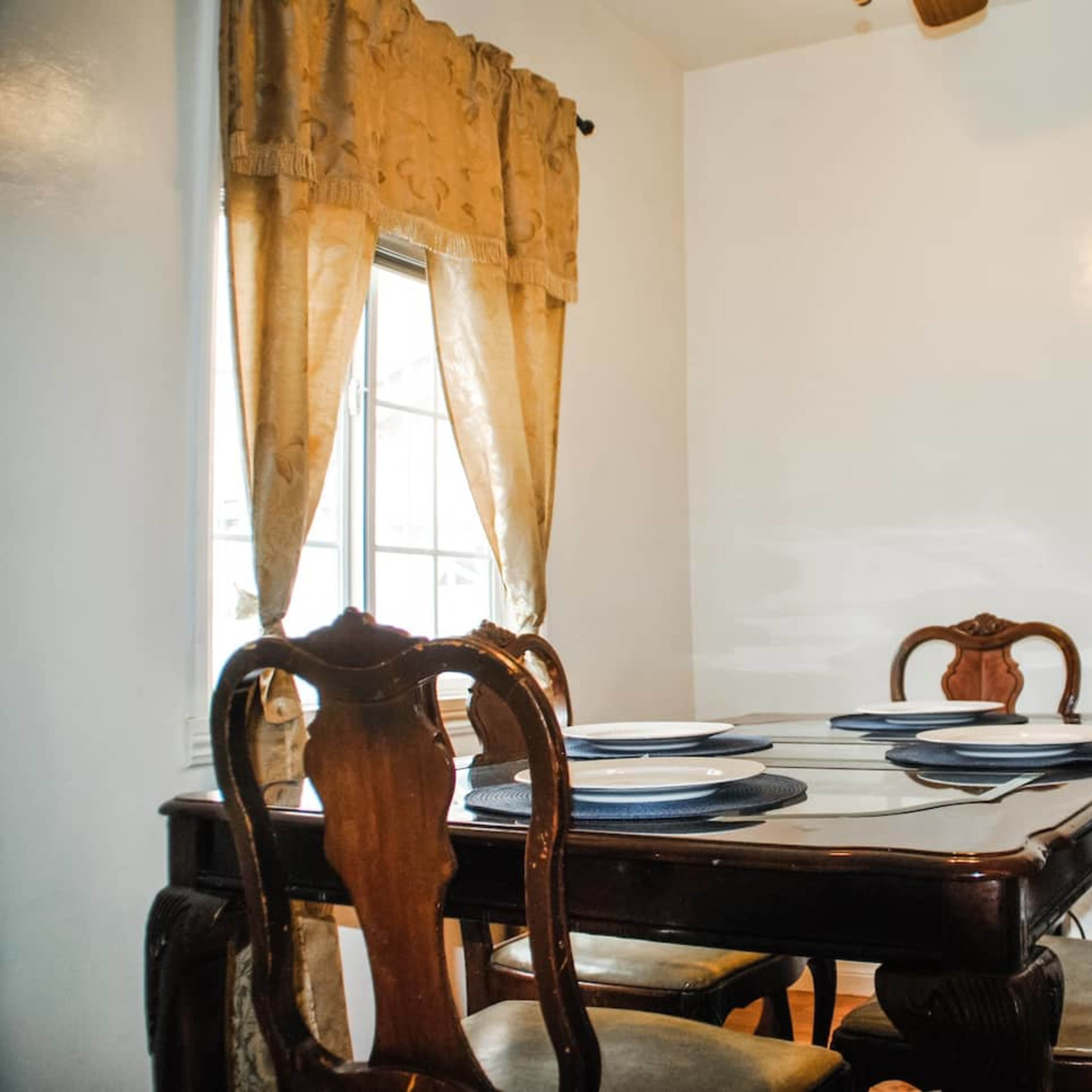 A wooden dining table set with plates is positioned near a window, alongside a water dispenser and a patterned curtain.
