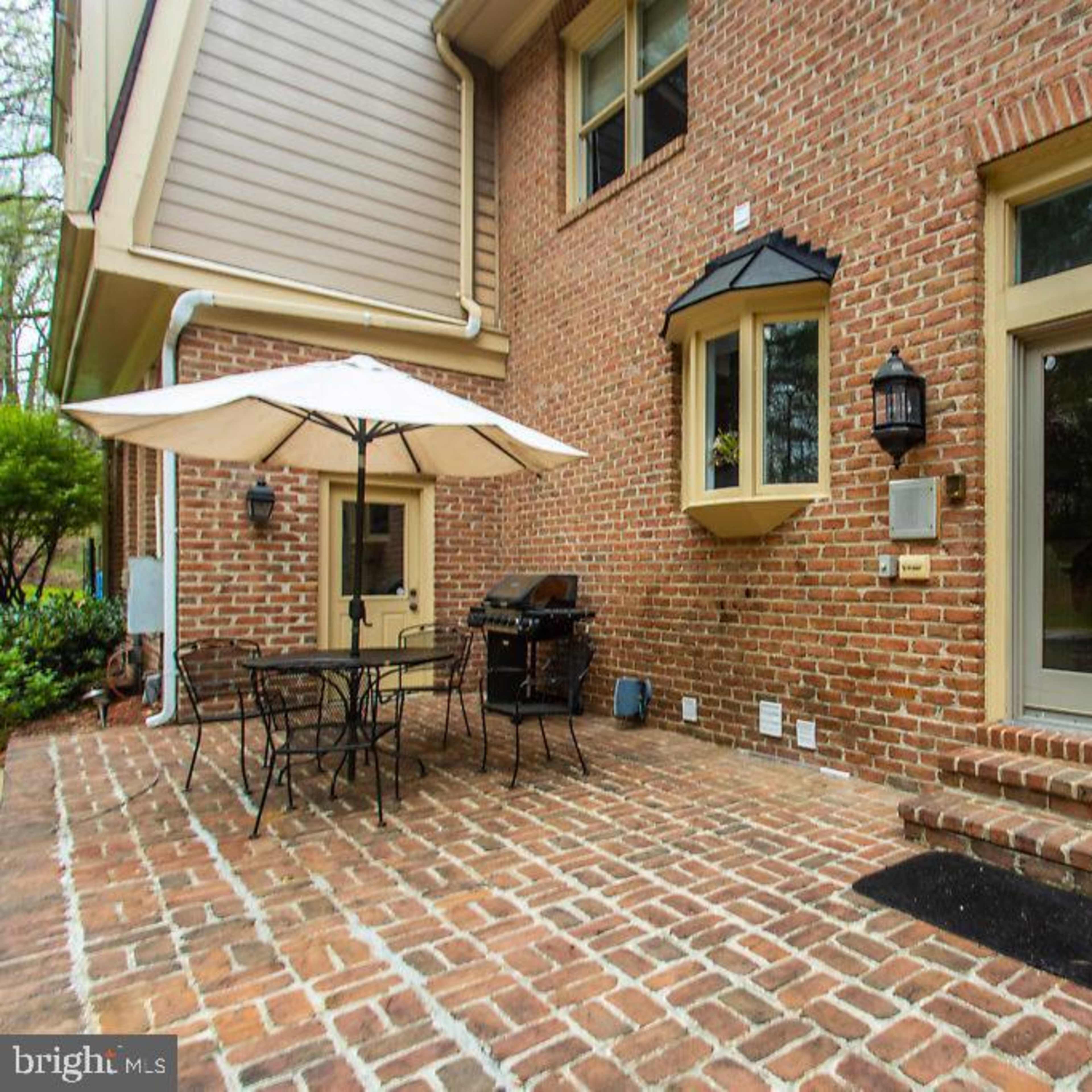 A brick patio with a table and chairs, an umbrella, and a grill, adjacent to a house with large glass doors.