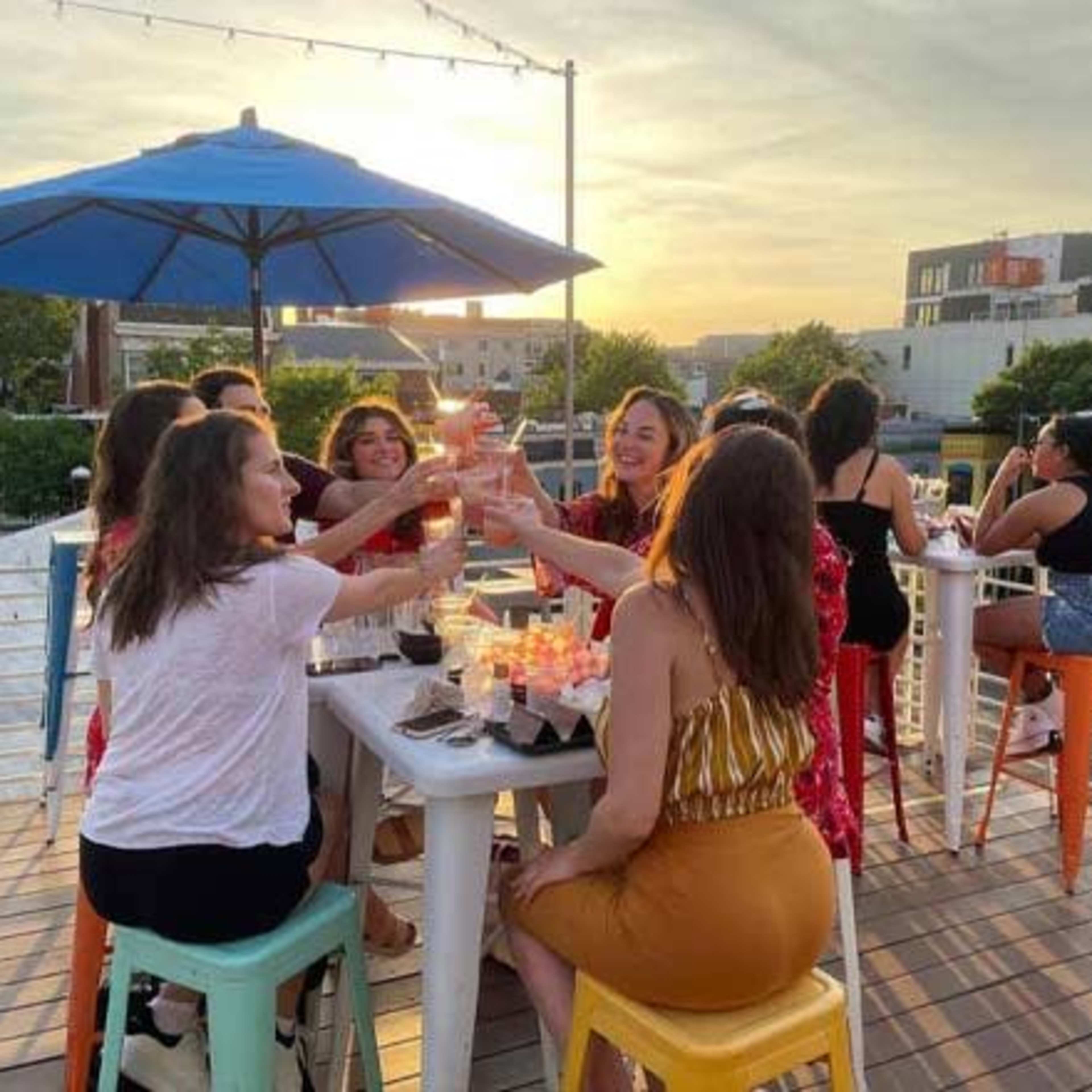 A group of people raise their glasses in a toast while seated at a table on a rooftop during sunset.