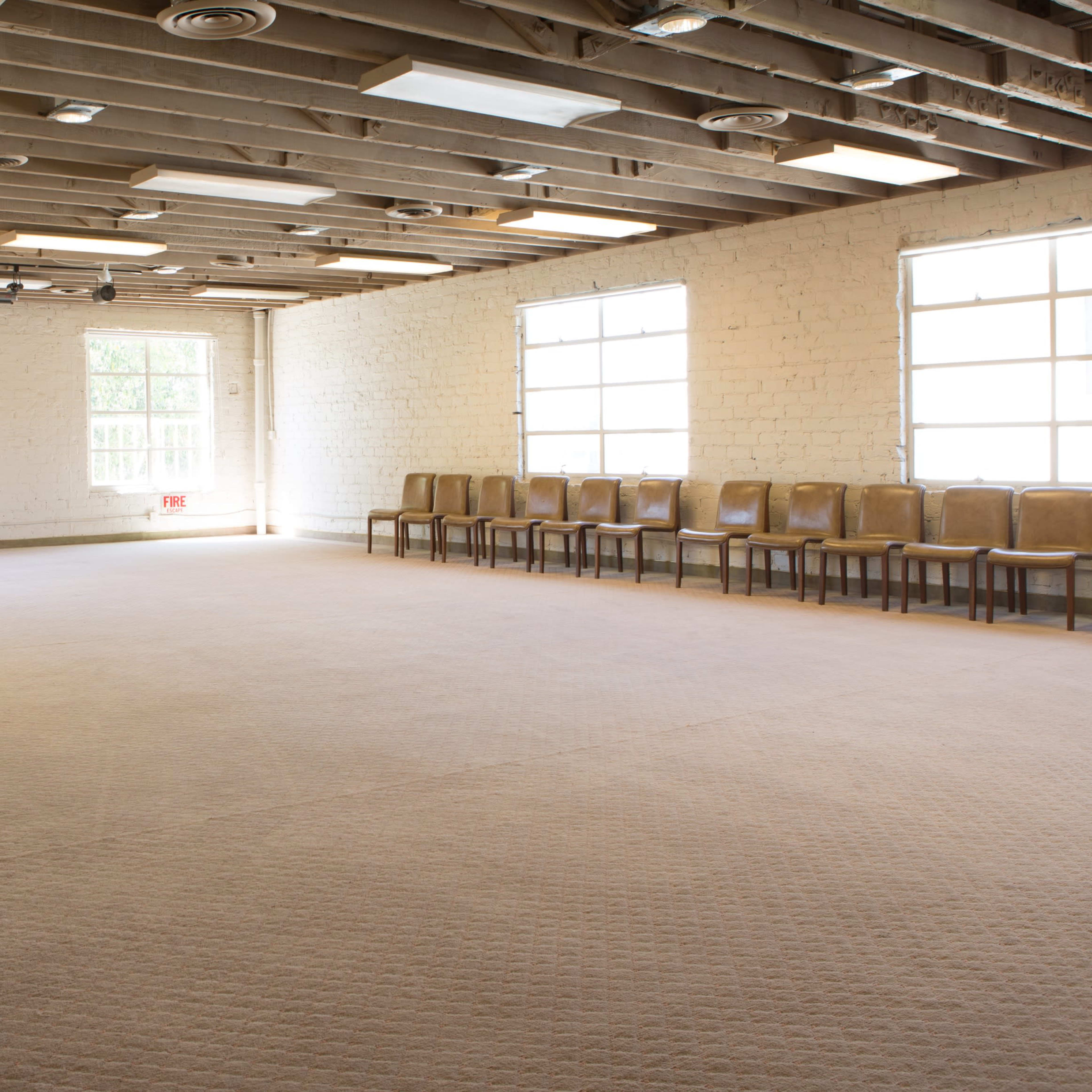 A spacious, empty room with a carpeted floor, wooden beams on the ceiling, and rows of chairs along one wall.