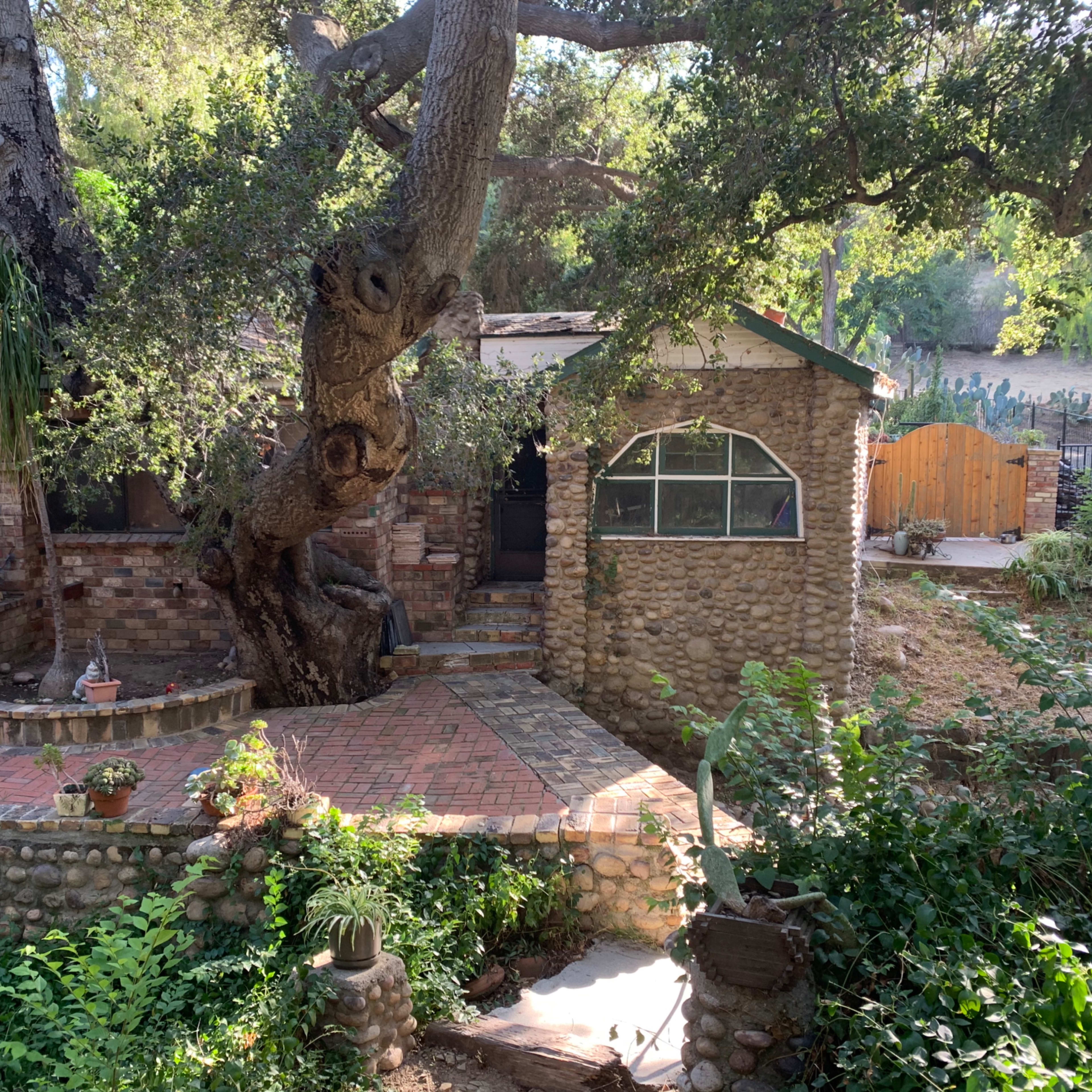 A rustic stone house with a large tree at its side and a winding brick pathway leads toward the entrance, surrounded by greenery.