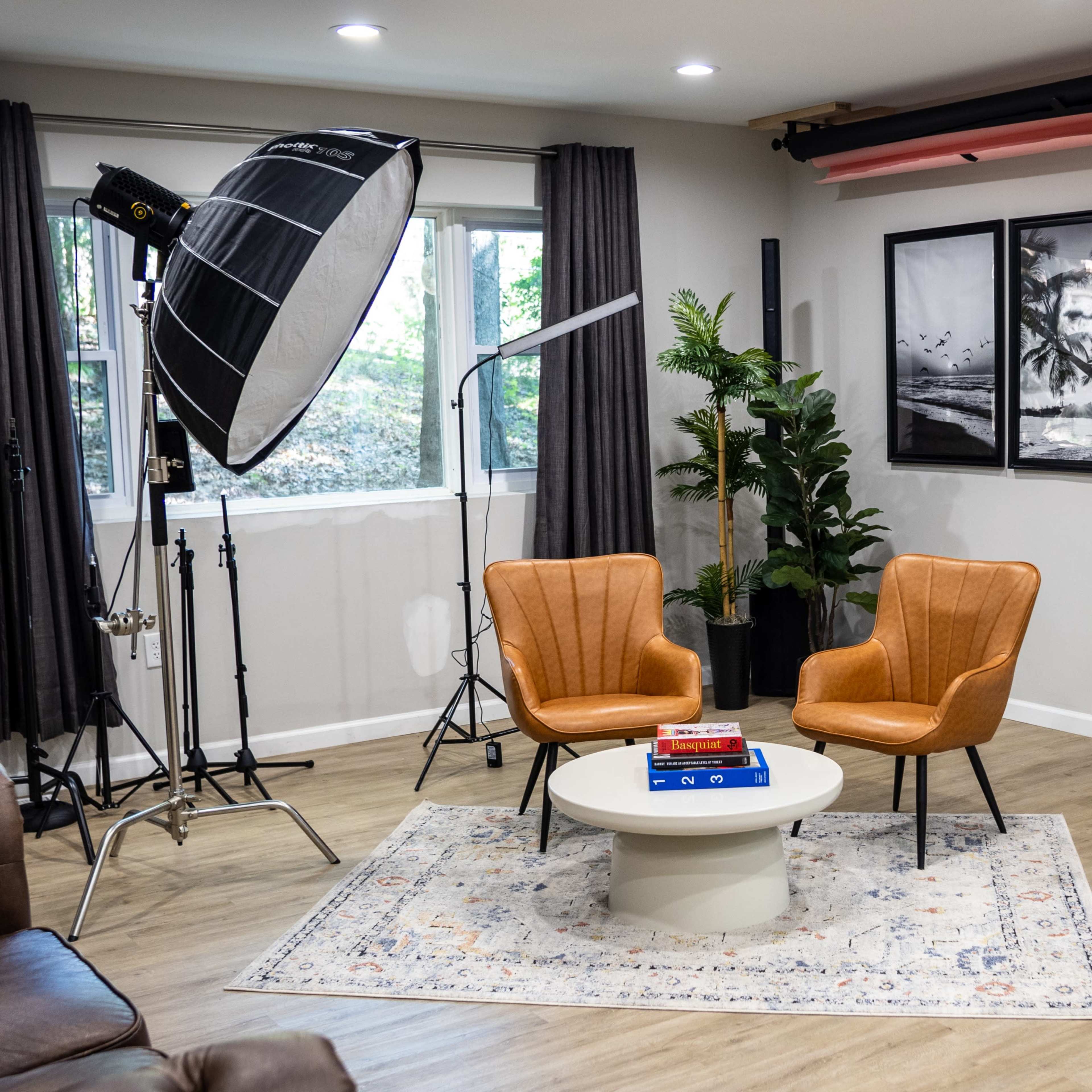 A modern studio setting with two orange chairs and a round table, surrounded by lighting equipment and framed photographs on the wall.