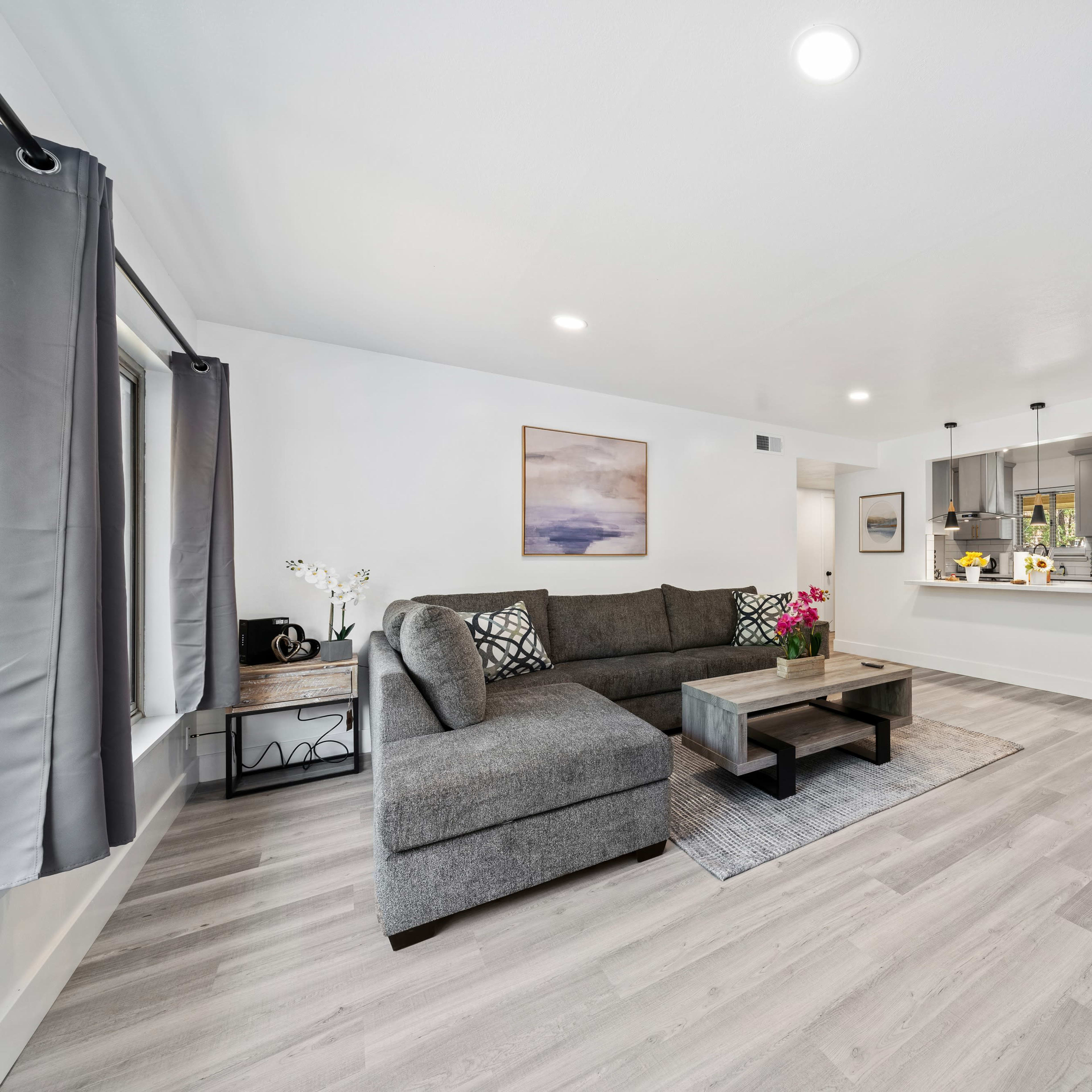 The spacious living room features a gray sectional sofa, a coffee table, and a view into a modern kitchen with bar seating and natural light.