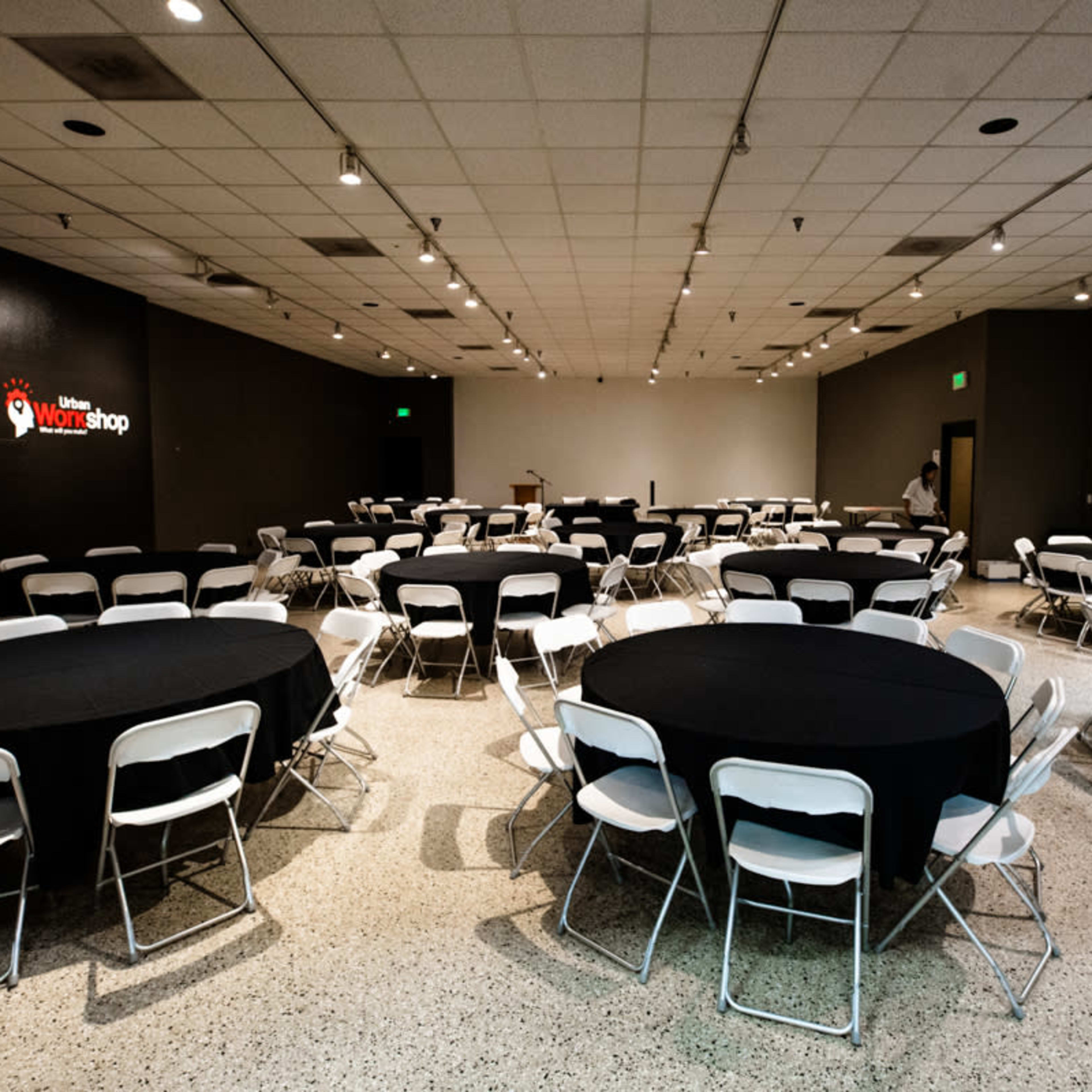 A large meeting room is arranged with multiple round tables covered in black cloth and surrounded by white chairs.