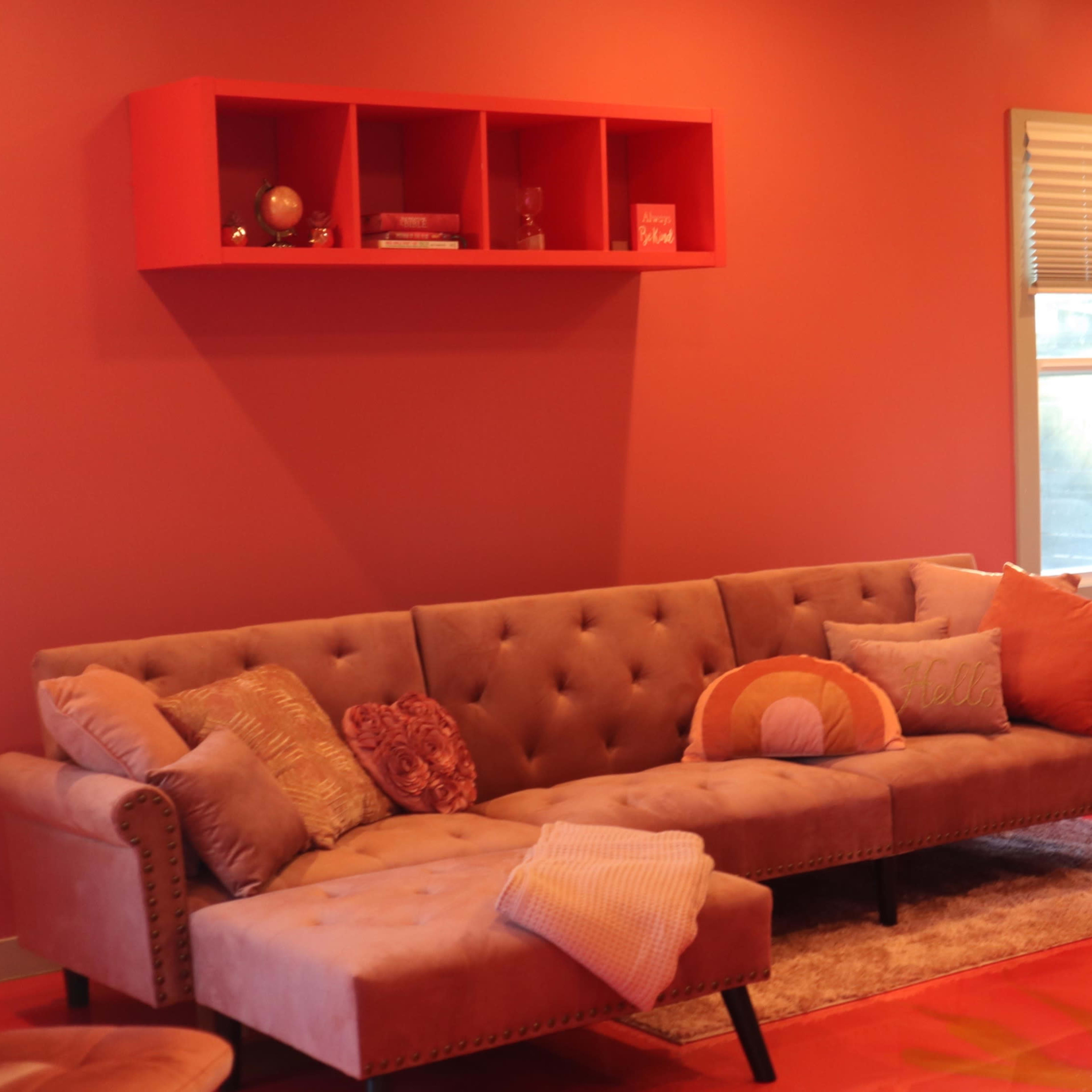 A living room with a pink wall, a gray tufted sofa, and a red shelf above it, accompanied by a small round table and bar stools in the background.