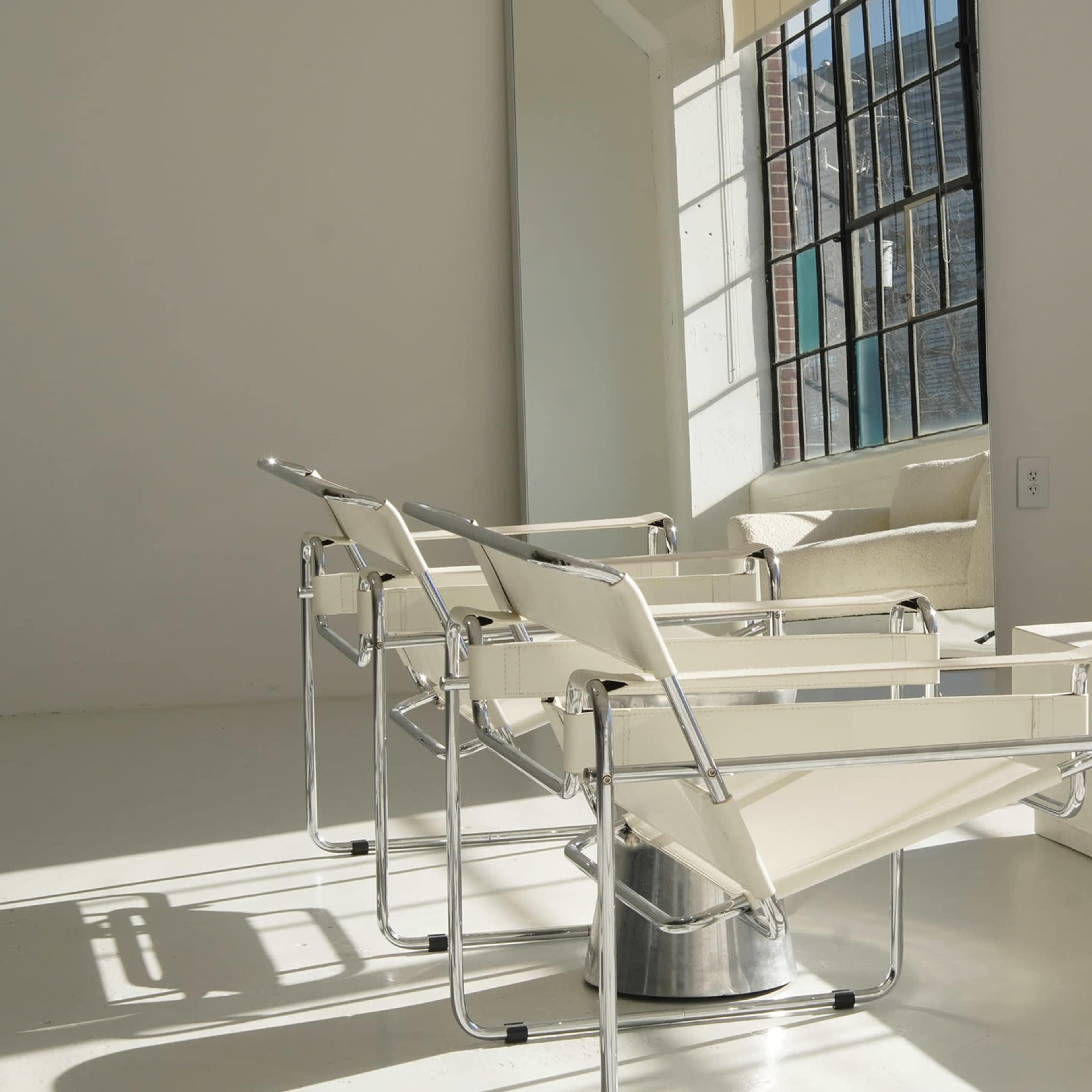 Two modern chairs facing a minimalist coffee table, with a large mirror reflecting light from a window in a bright interior space.