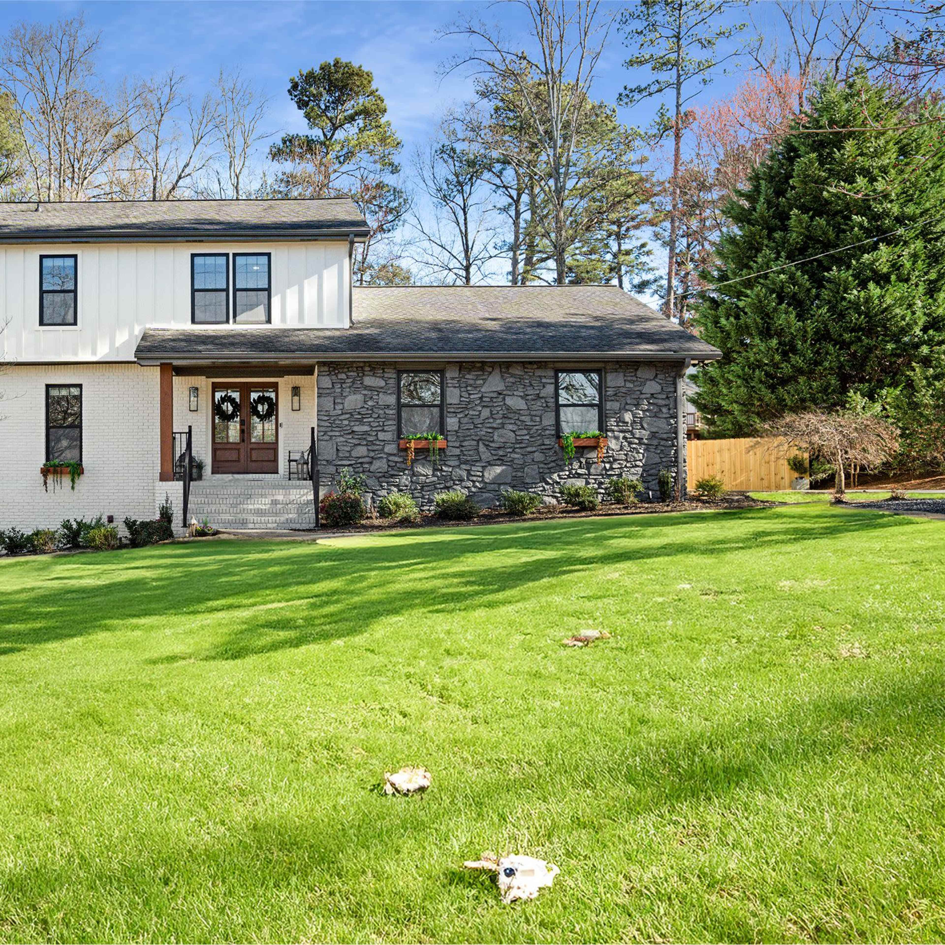 A two-story house featuring a combination of white exterior siding and stone accents, situated on a grassy lawn with trees in the background.