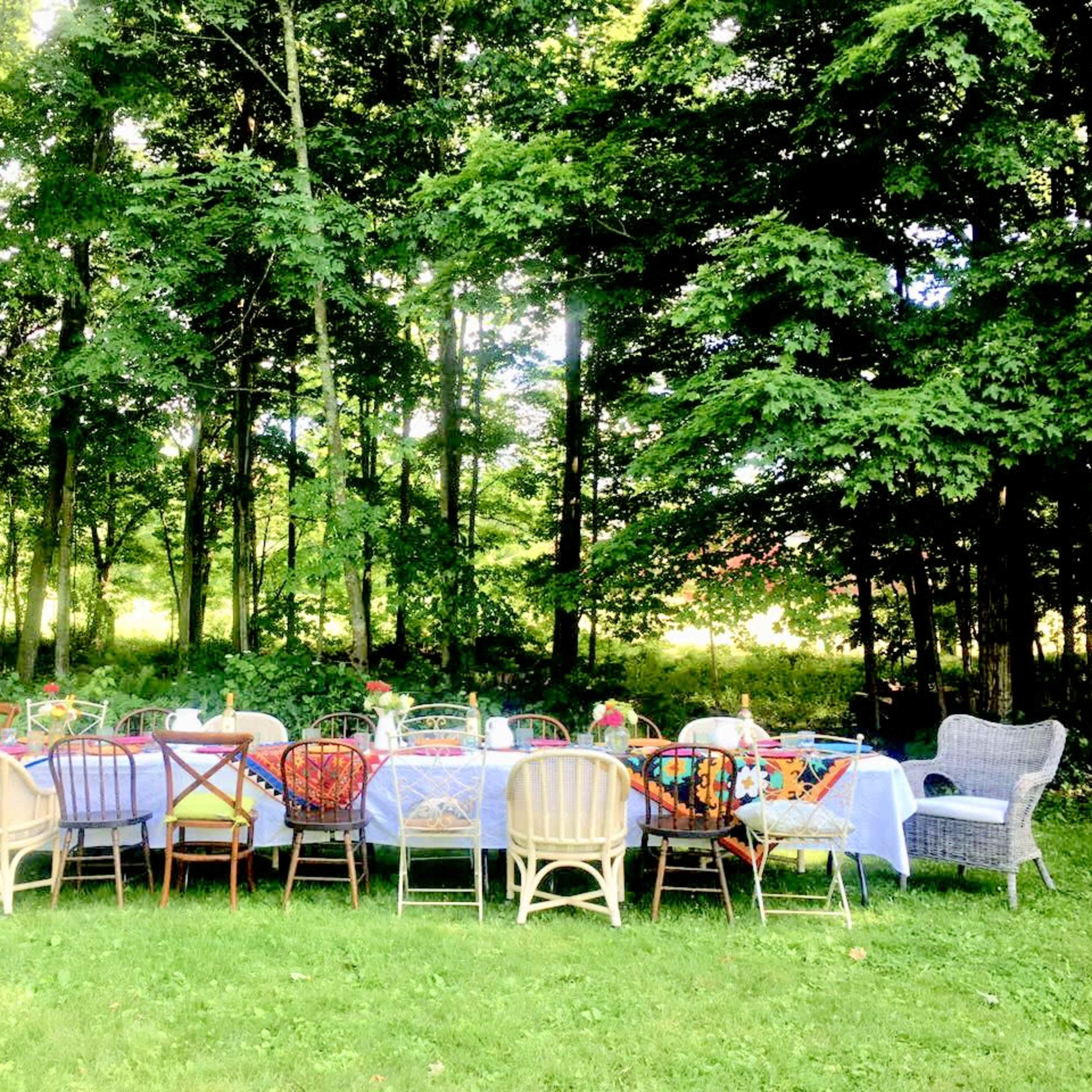 A long table set for a meal is surrounded by various chairs in a wooded outdoor setting.