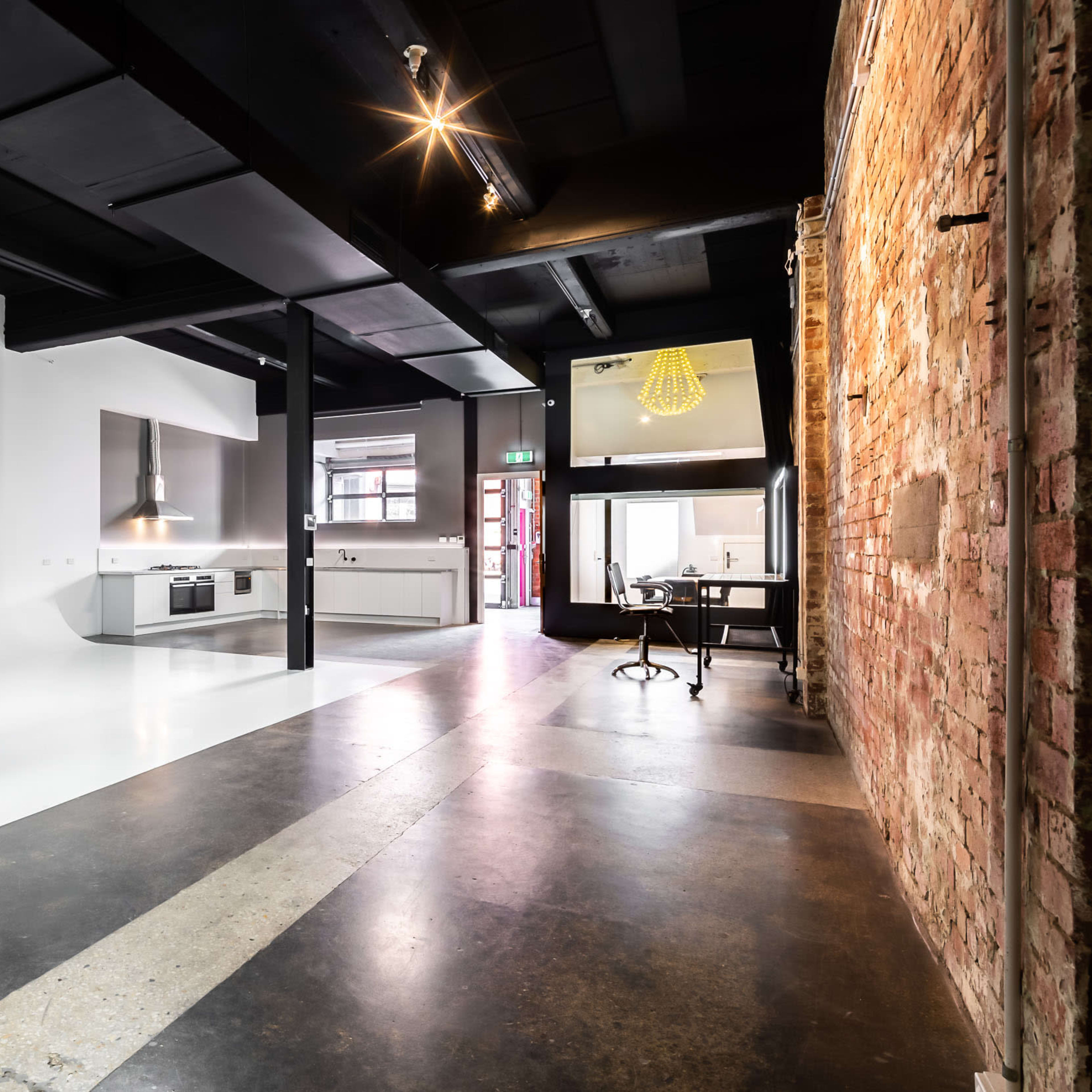 A modern, open-plan space features a kitchen area with sleek appliances, exposed brick walls, and a combination of polished concrete and white flooring.