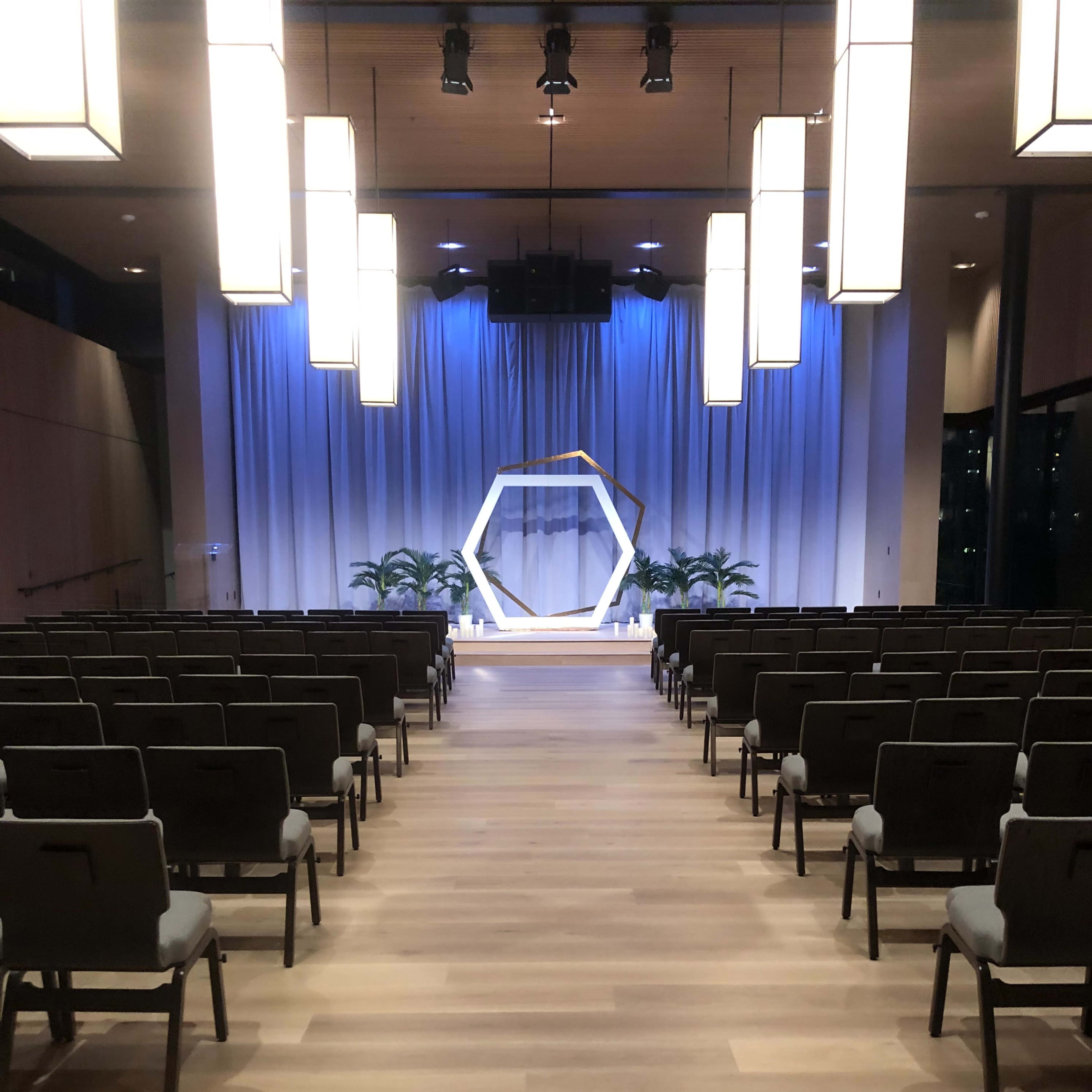 A modern auditorium with rows of chairs facing a stage featuring a hexagonal backdrop and decorative plants.