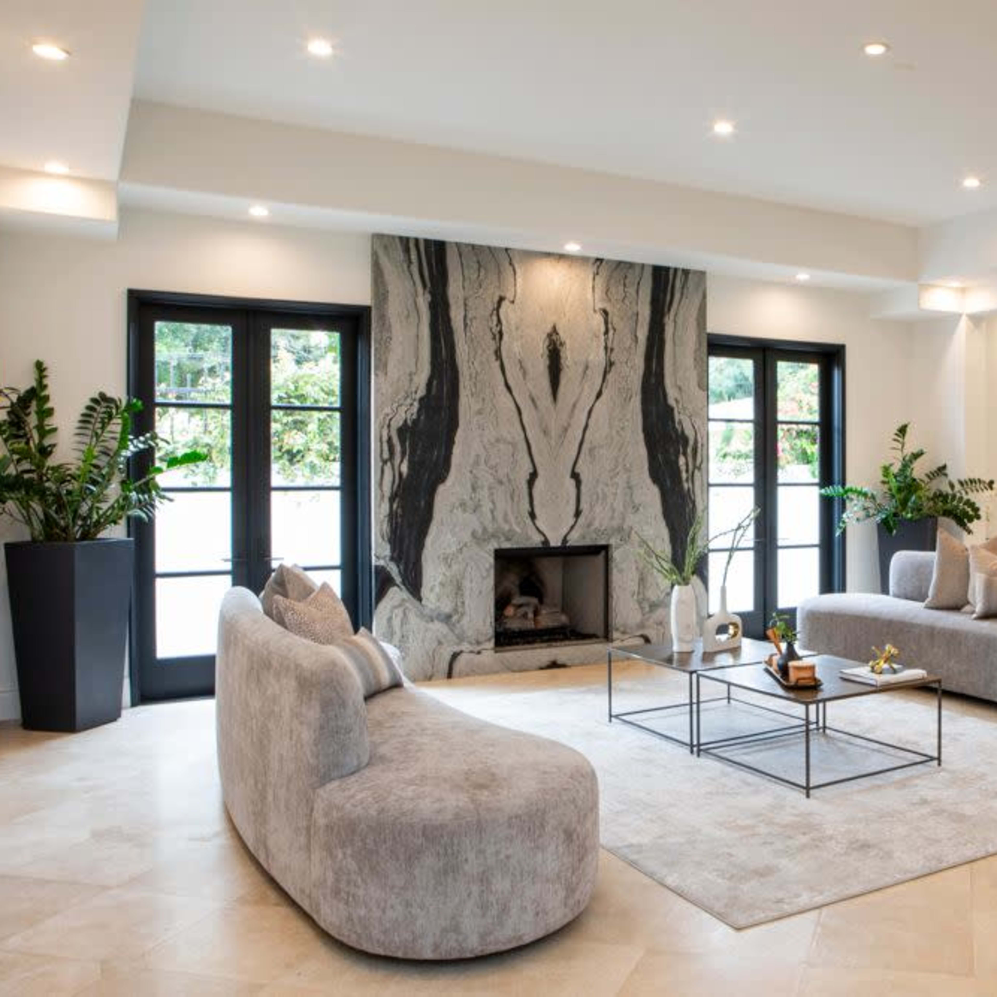 A modern living room features a large, curved gray sofa, a matching loveseat, a black coffee table, a stone fireplace, and large windows surrounded by potted plants.