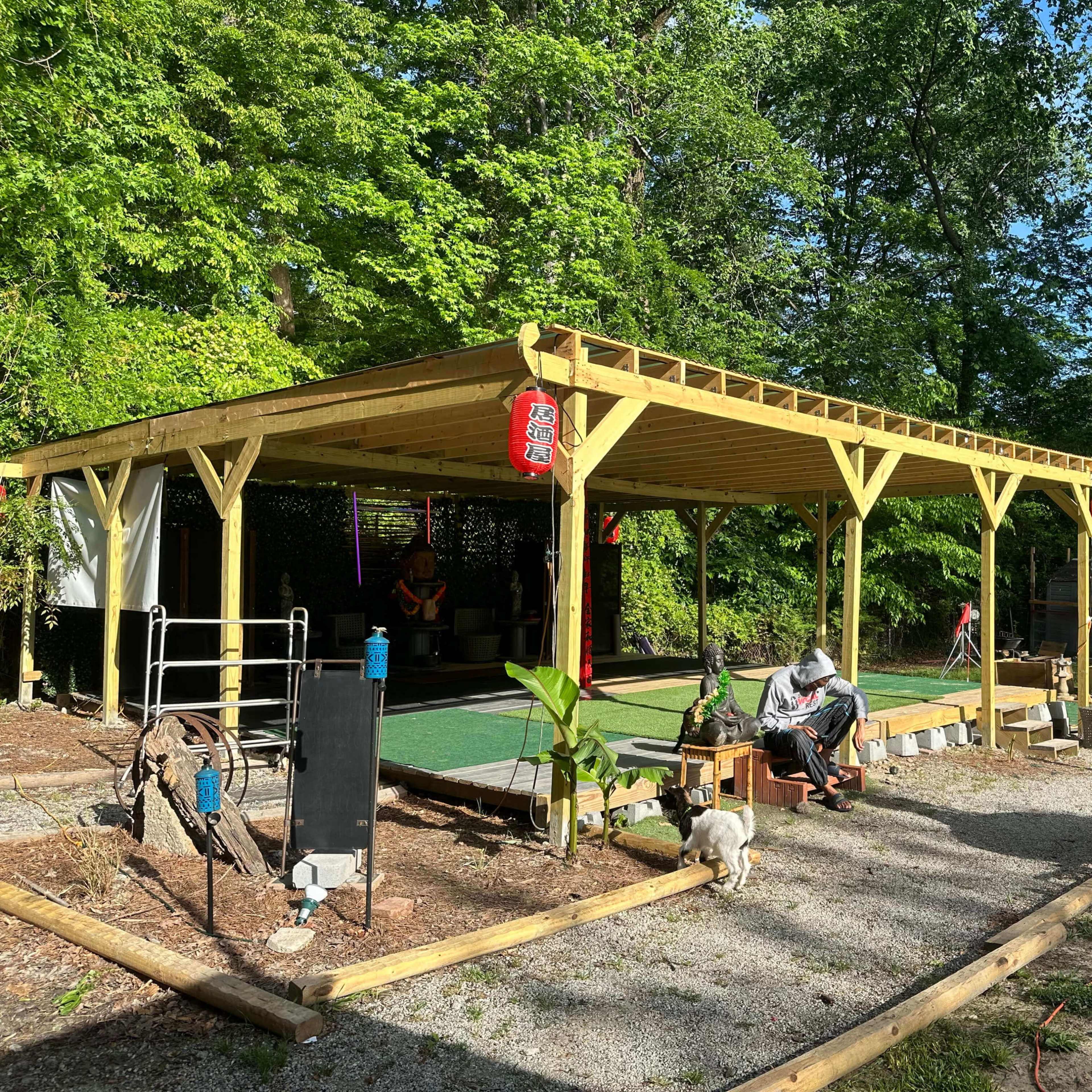 A wooden gazebo structure with benches and a stone pathway is surrounded by green trees and garden elements.
