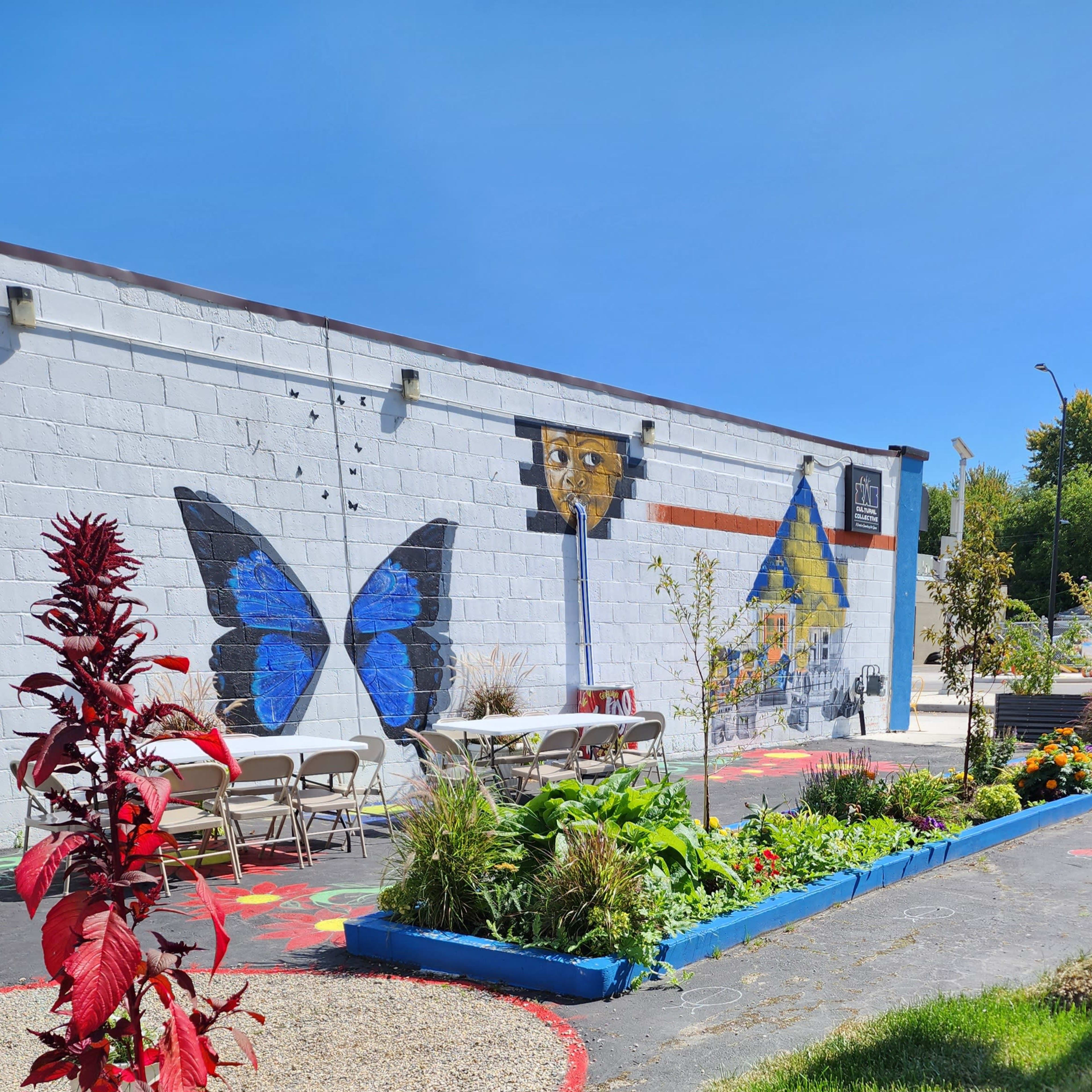 A colorful mural featuring a butterfly and a house decorates a wall in a garden setting with tables and greenery.