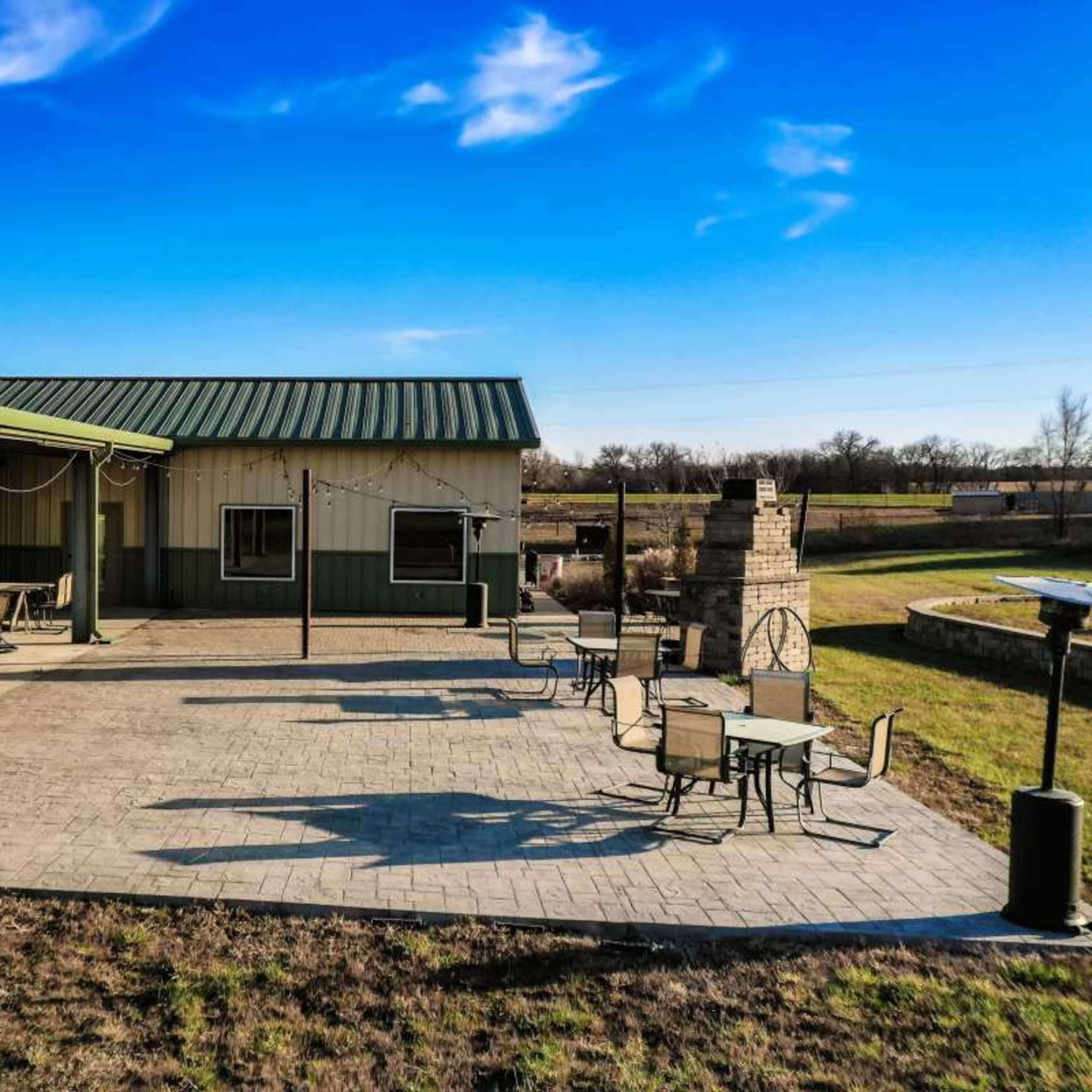 A spacious outdoor patio area with several tables and chairs, adjacent to a metal building and a stone fireplace.