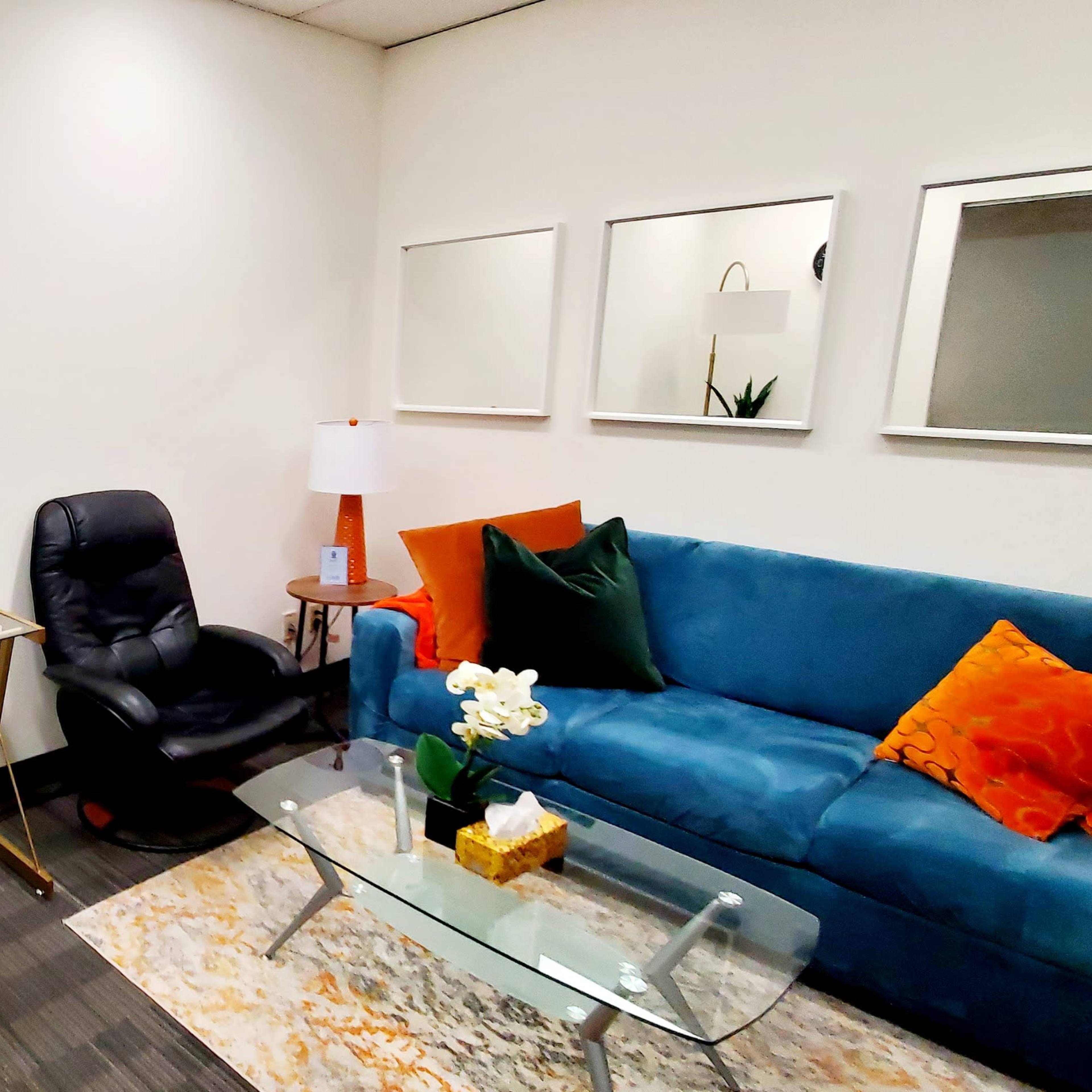 A small office space with a blue couch, a glass coffee table, a desk with a lamp, and a black chair.