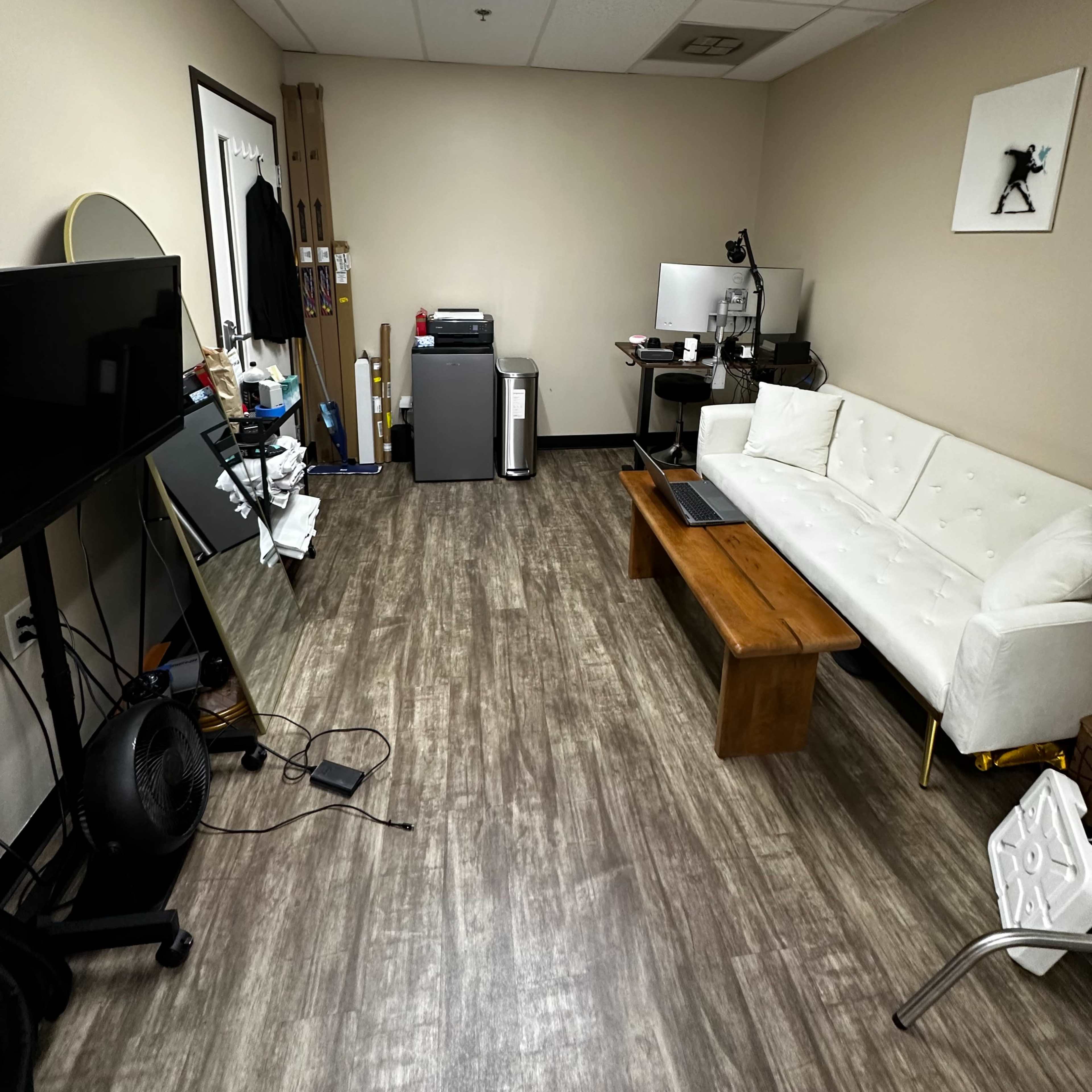 An office space with a white sofa, a wooden coffee table, a television, and a computer setup against a light-colored wall.