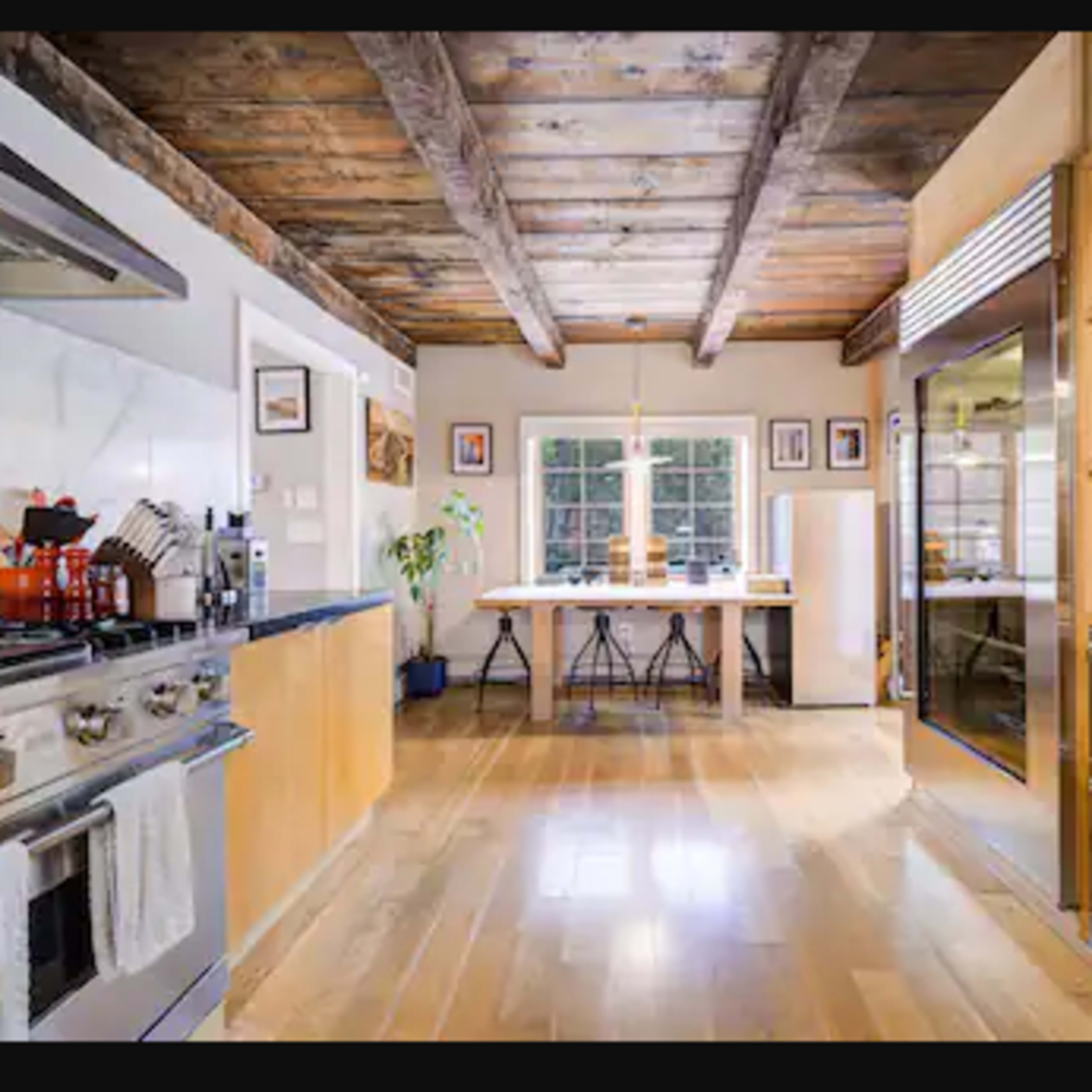 A modern kitchen features stainless steel appliances, wooden cabinetry, and a central dining table illuminated by natural light from a large window.