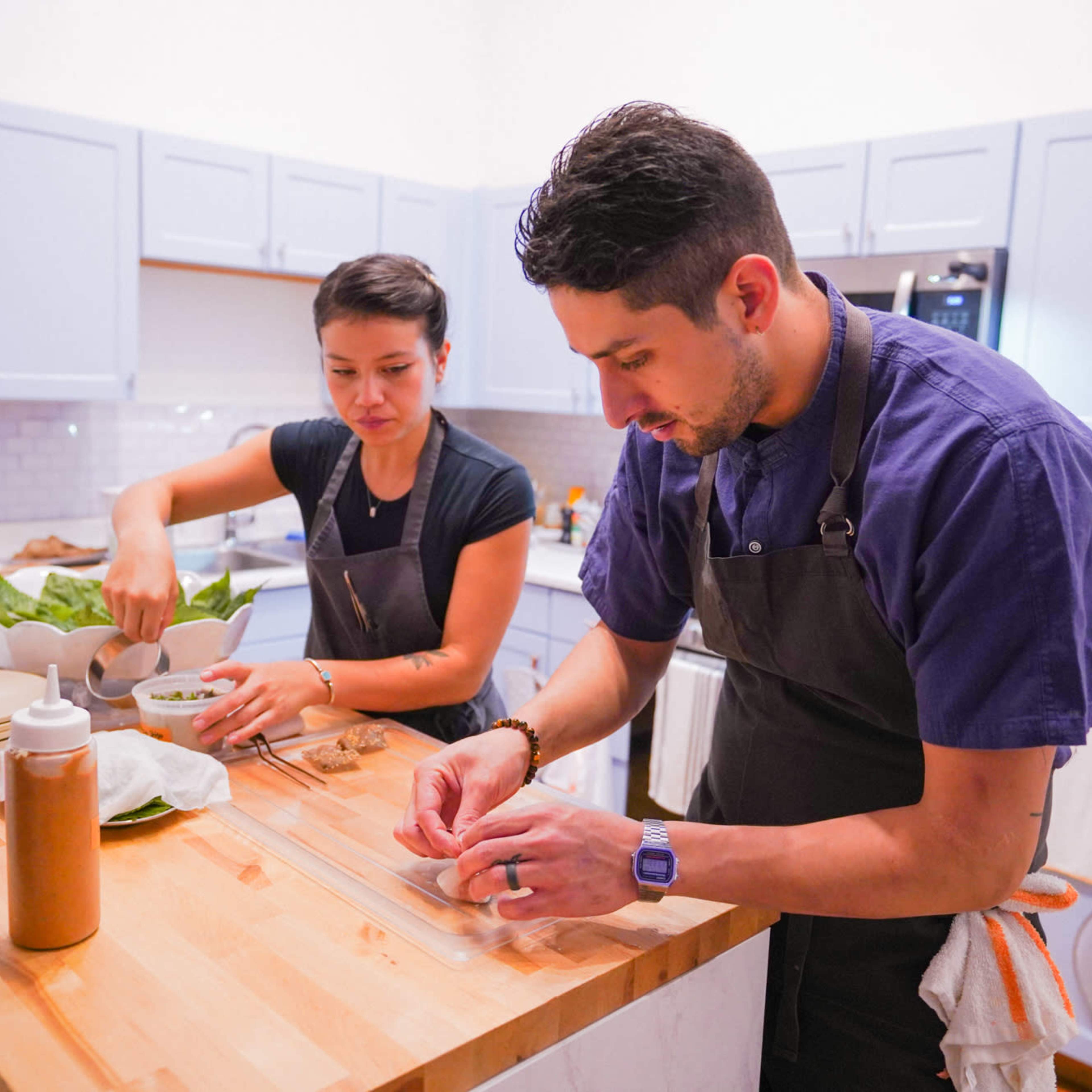 Two chefs are preparing ingredients in a kitchen, with one chopping vegetables and the other assembling a dish on a wooden countertop.