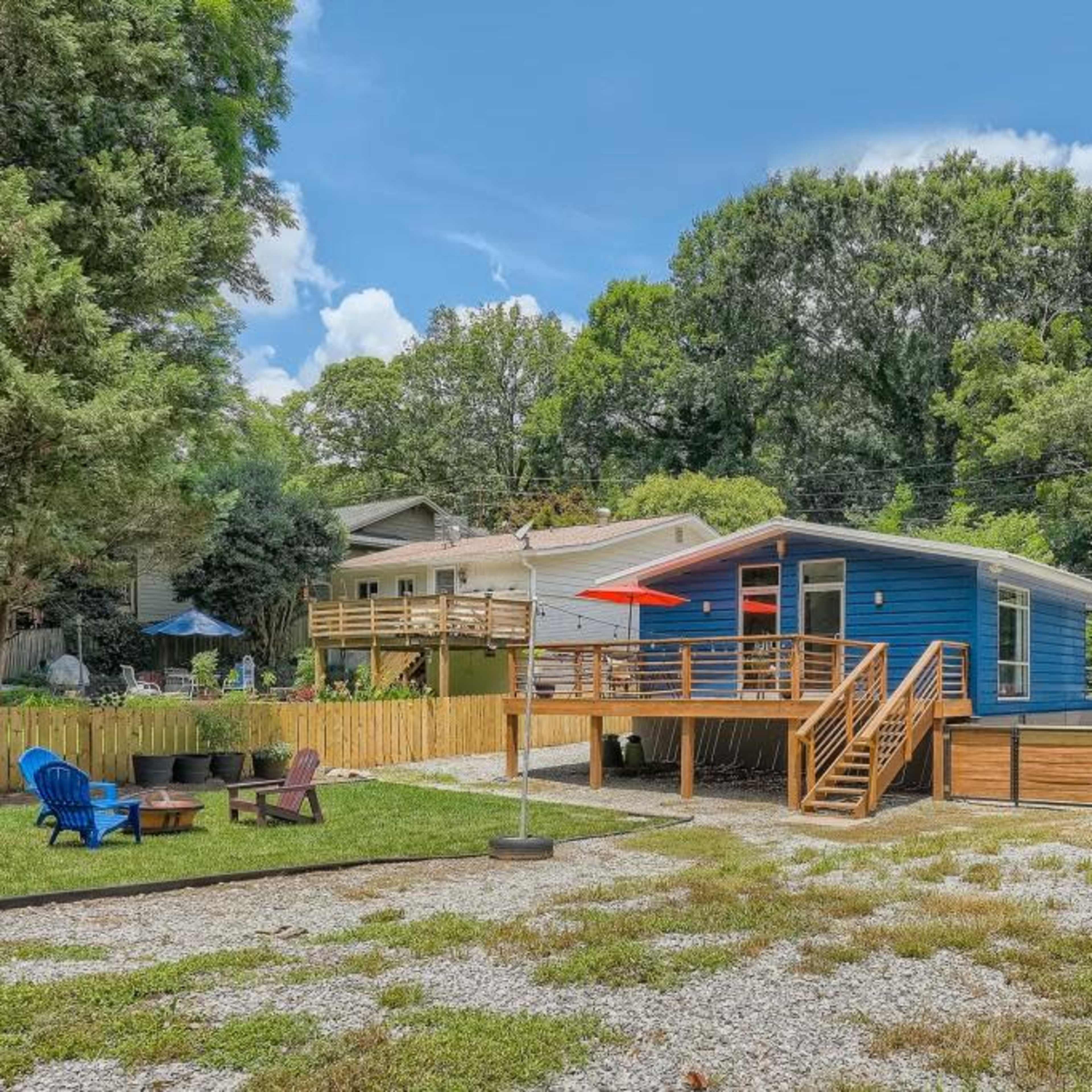 A blue house with a wooden deck and red umbrella is set in a grassy yard surrounded by trees and a gravel area with outdoor seating.