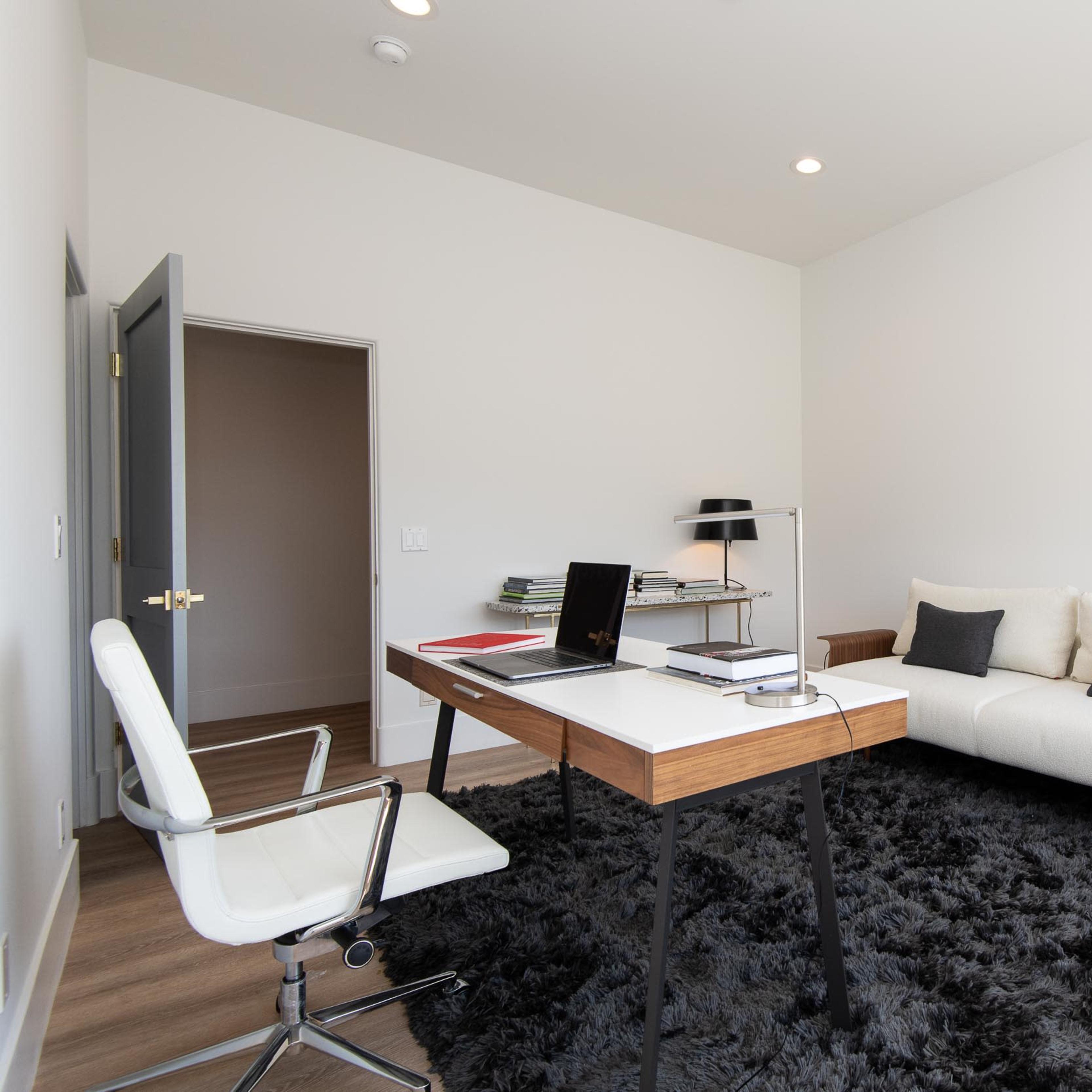 A modern office space features a wooden desk with a laptop and books, a white couch, and a black rug on a hardwood floor.