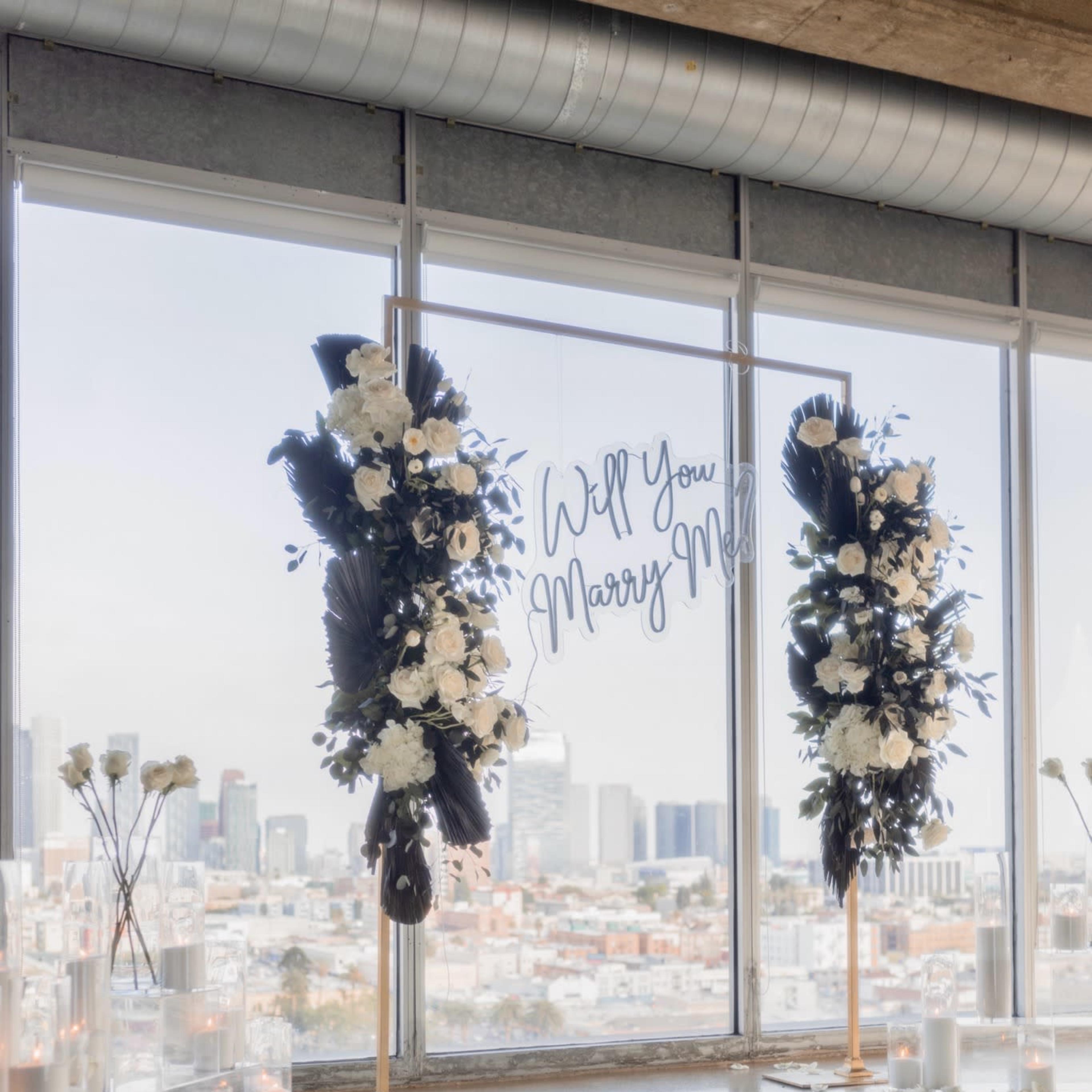 A romantic proposal setup features a large floral display on either side of a glass window with a view of the city, complemented by a carpet of rose petals and candles.