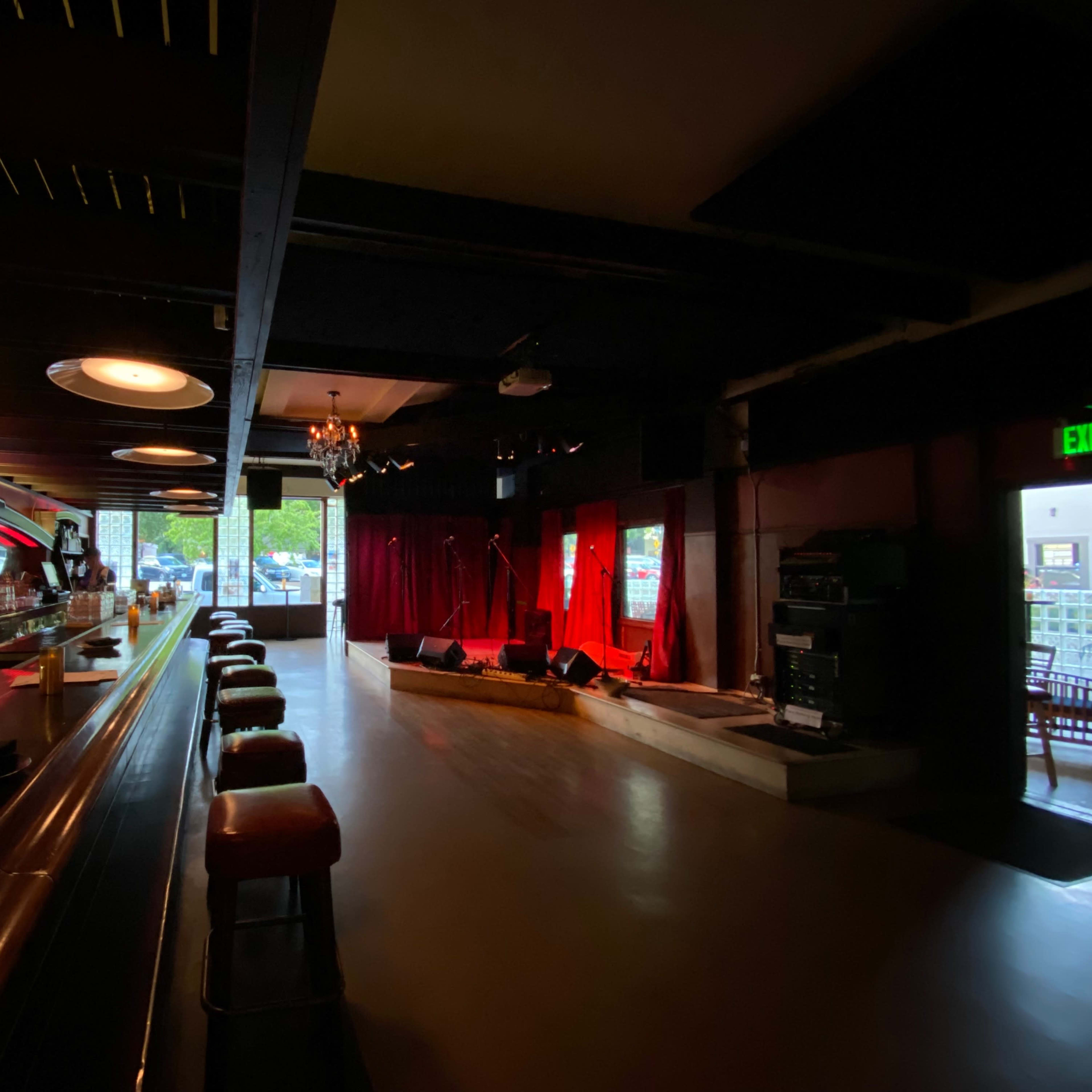 A dimly lit bar with a long counter and several stools facing a small stage with red curtains, illuminated by soft lighting.