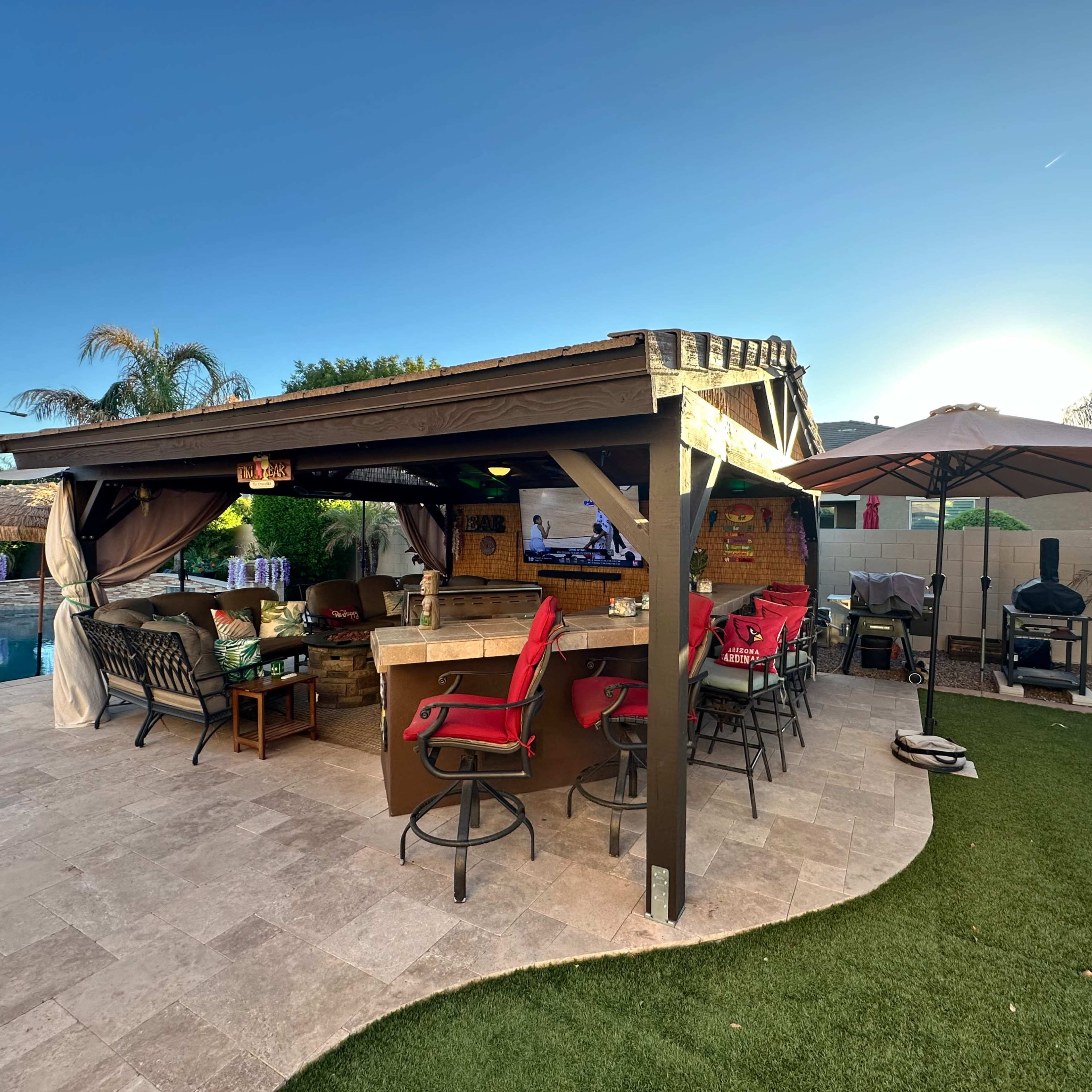 A covered outdoor space features a bar area with red chairs, nearby seating, and a pool in the background.