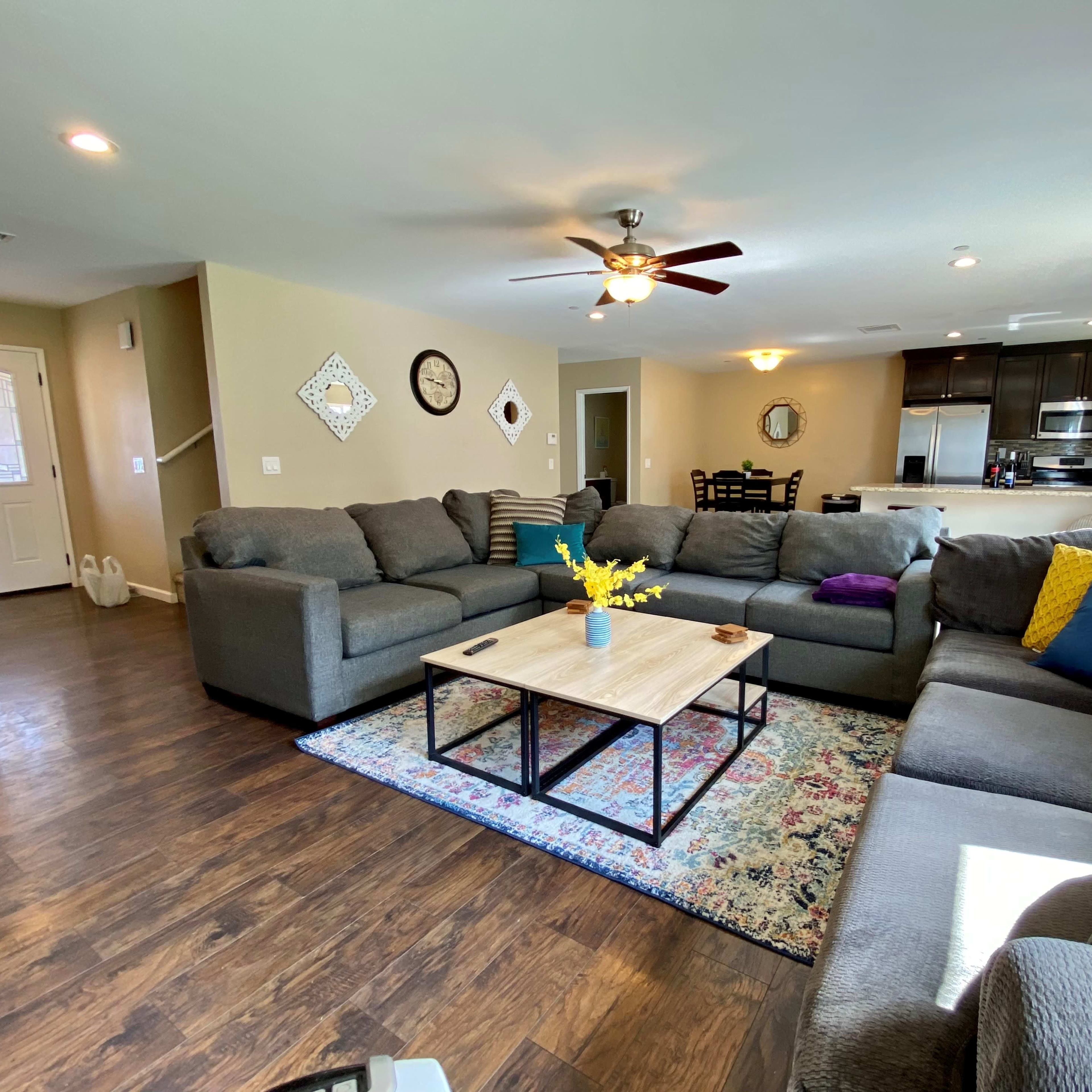 The living room features a large sectional sofa arranged around a coffee table, with a colorful area rug and a view into the kitchen and dining area.