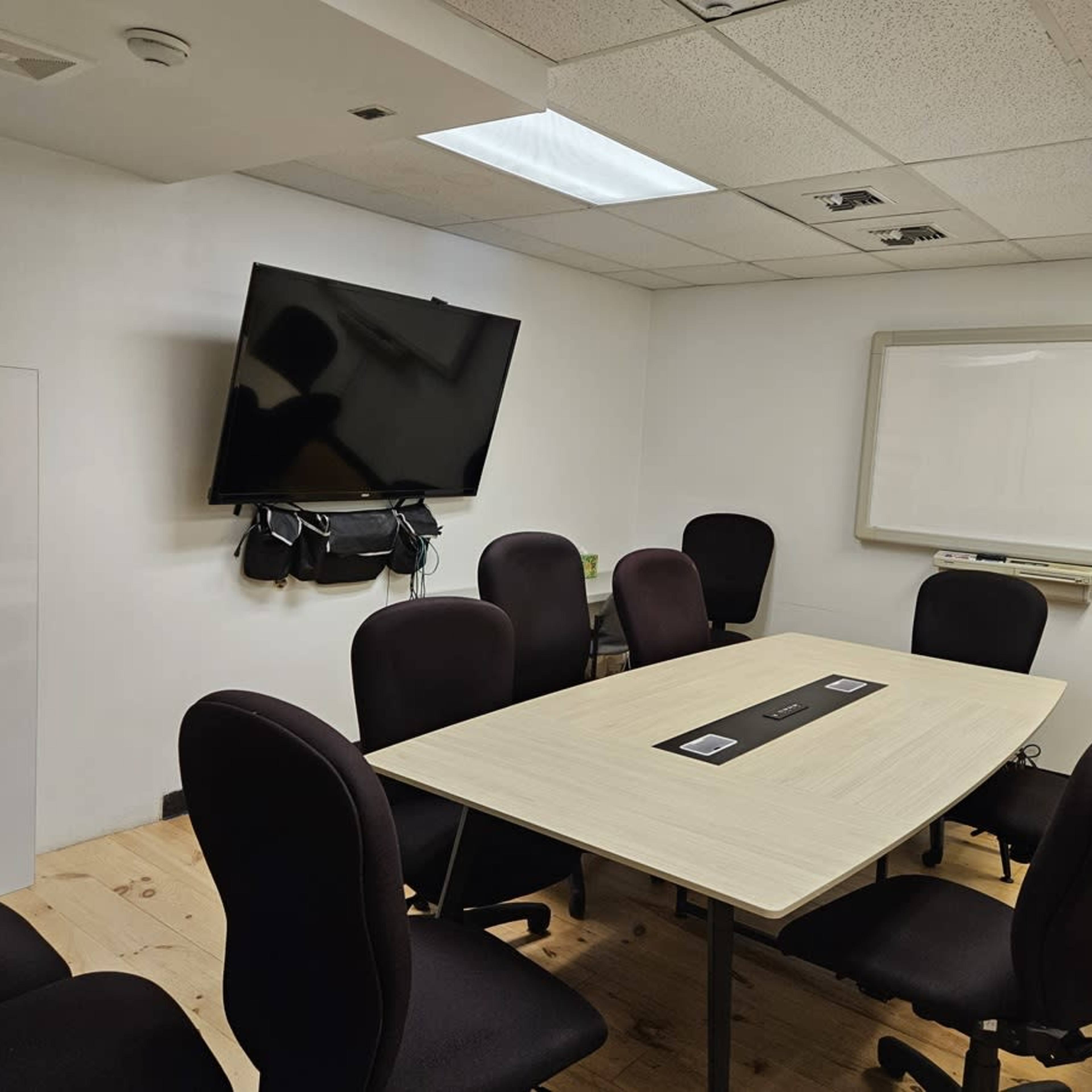 A conference room features a large table surrounded by eight black chairs, a wall-mounted TV, and a whiteboard.