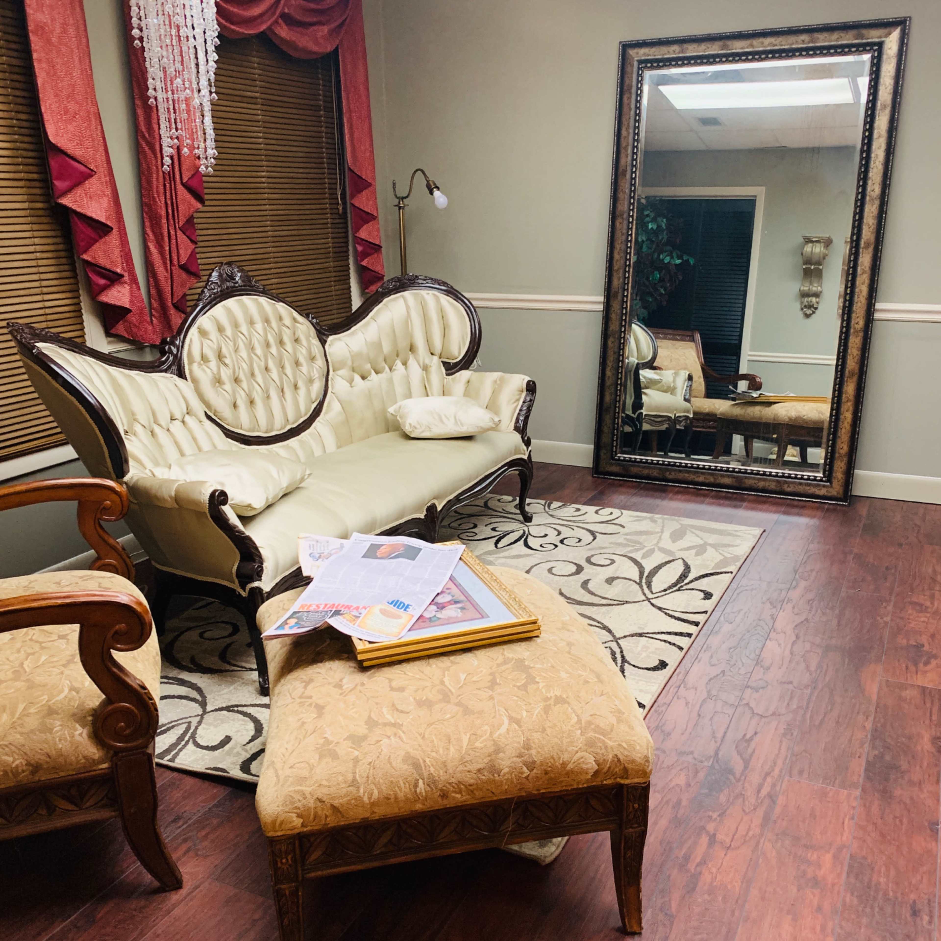 A furnished room with a vintage couch, an ottoman, and a large mirror, featuring a patterned rug and wooden flooring.