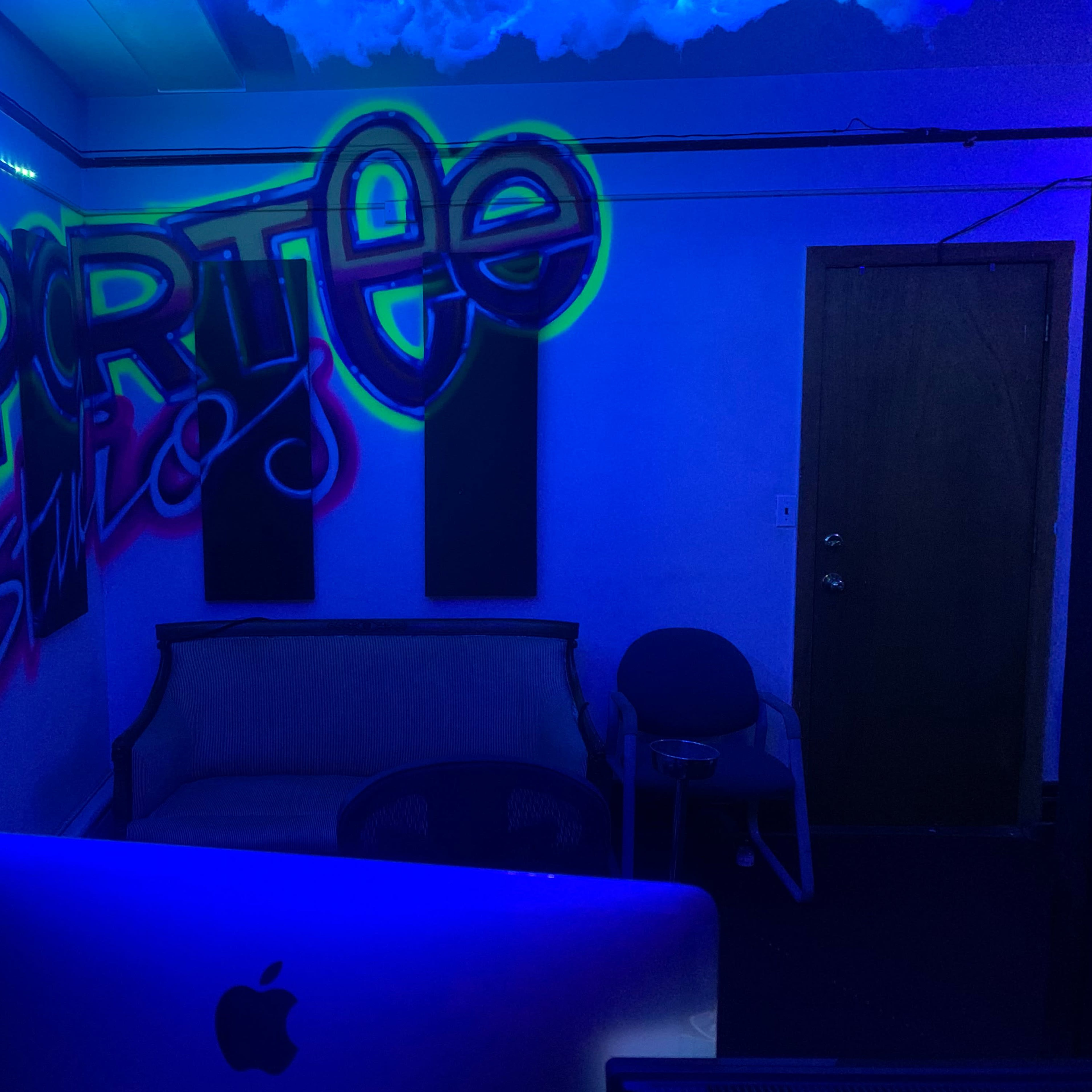 A dimly lit room featuring a graffiti mural that reads "Daprttee Studios" on one wall, with a couch and a chair visible in the foreground.
