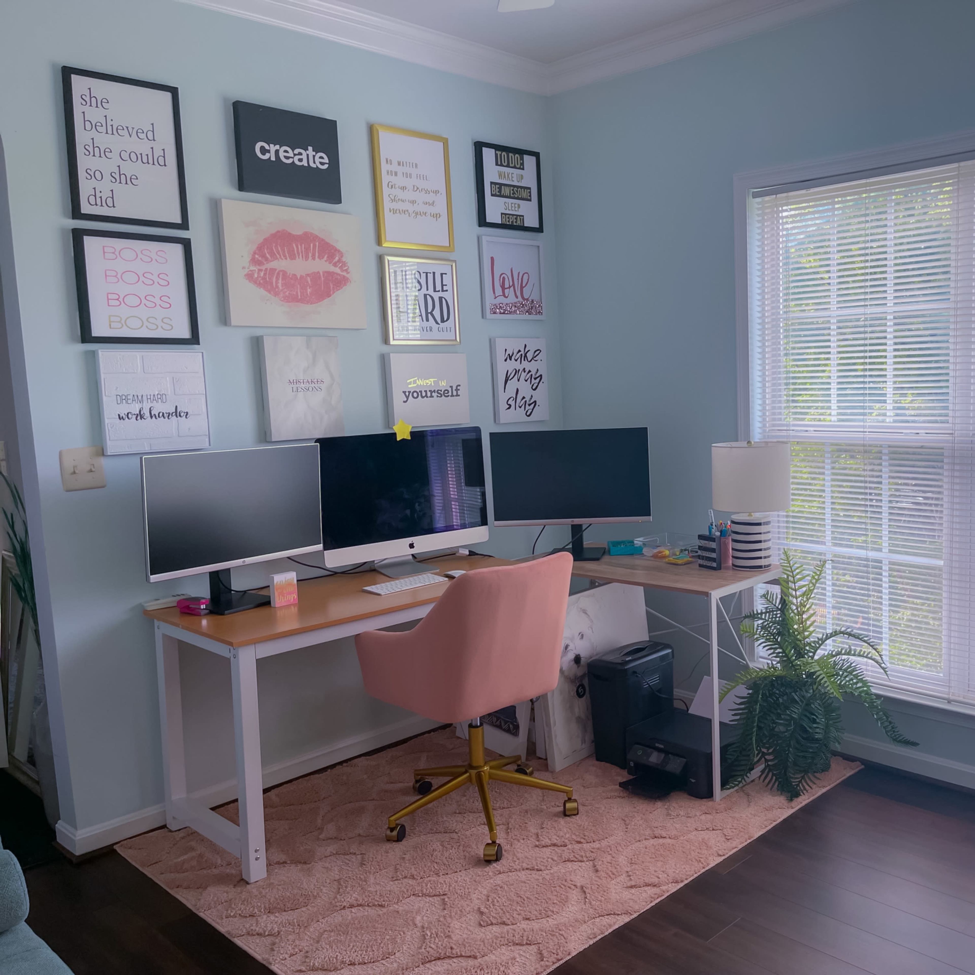 A home office features a desk with dual monitors, a pink chair, and inspirational wall art, along with a large window that brings in natural light.
