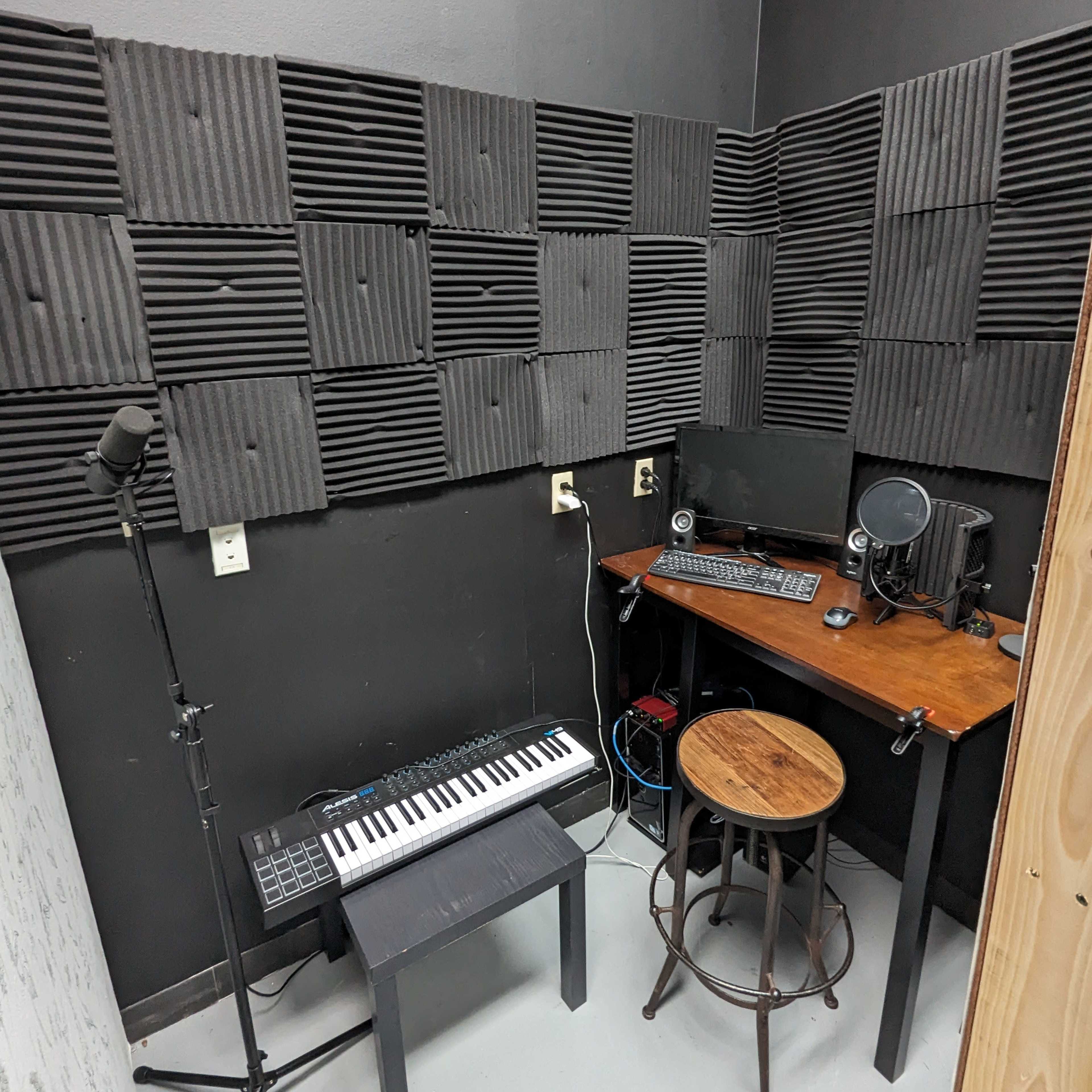 A small soundproof studio with acoustic foam panels on the walls, a wooden desk with a computer and microphone, and a keyboard situated next to a stool.