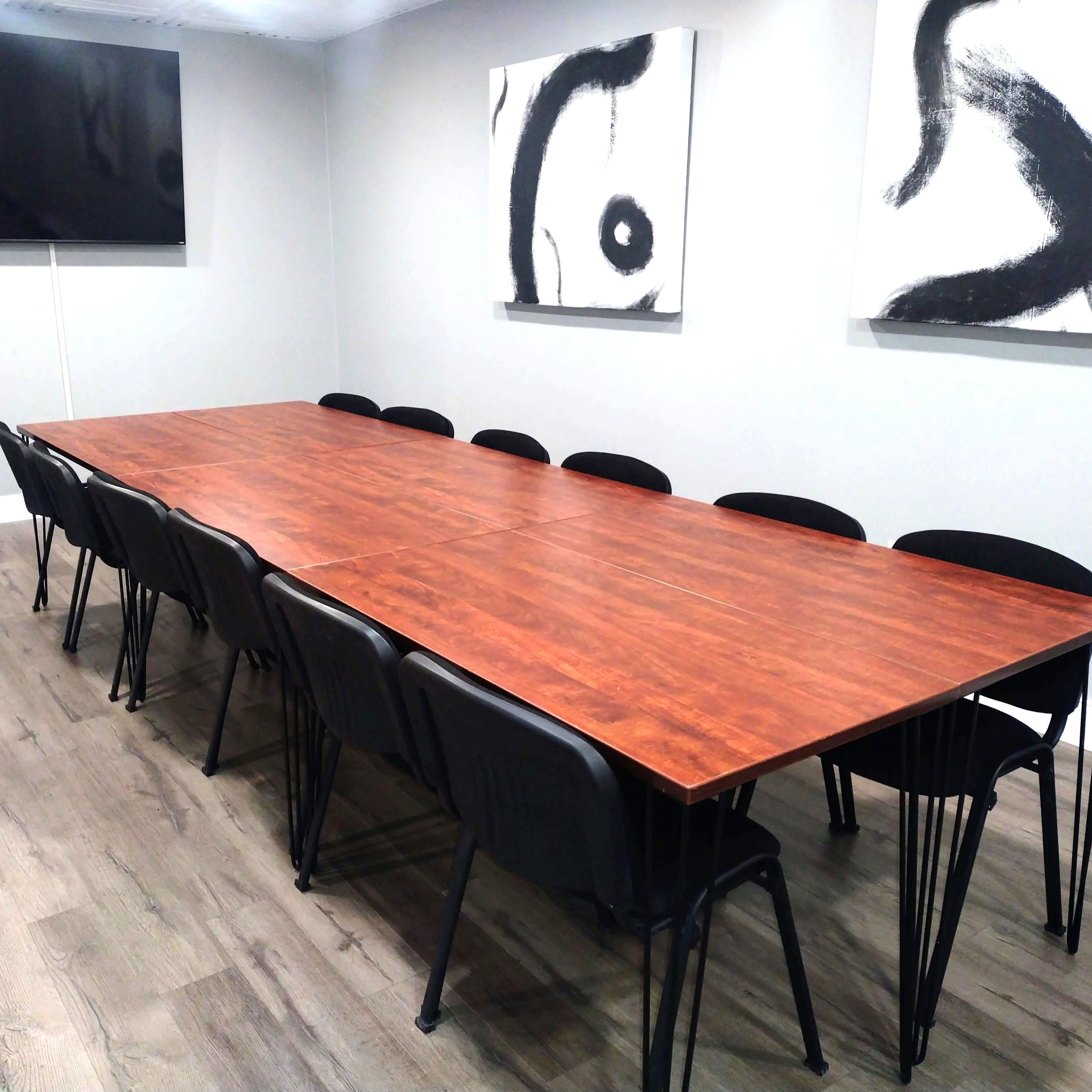 A long wooden table with black chairs is set in a room featuring abstract art on the walls and a television mounted on one side.