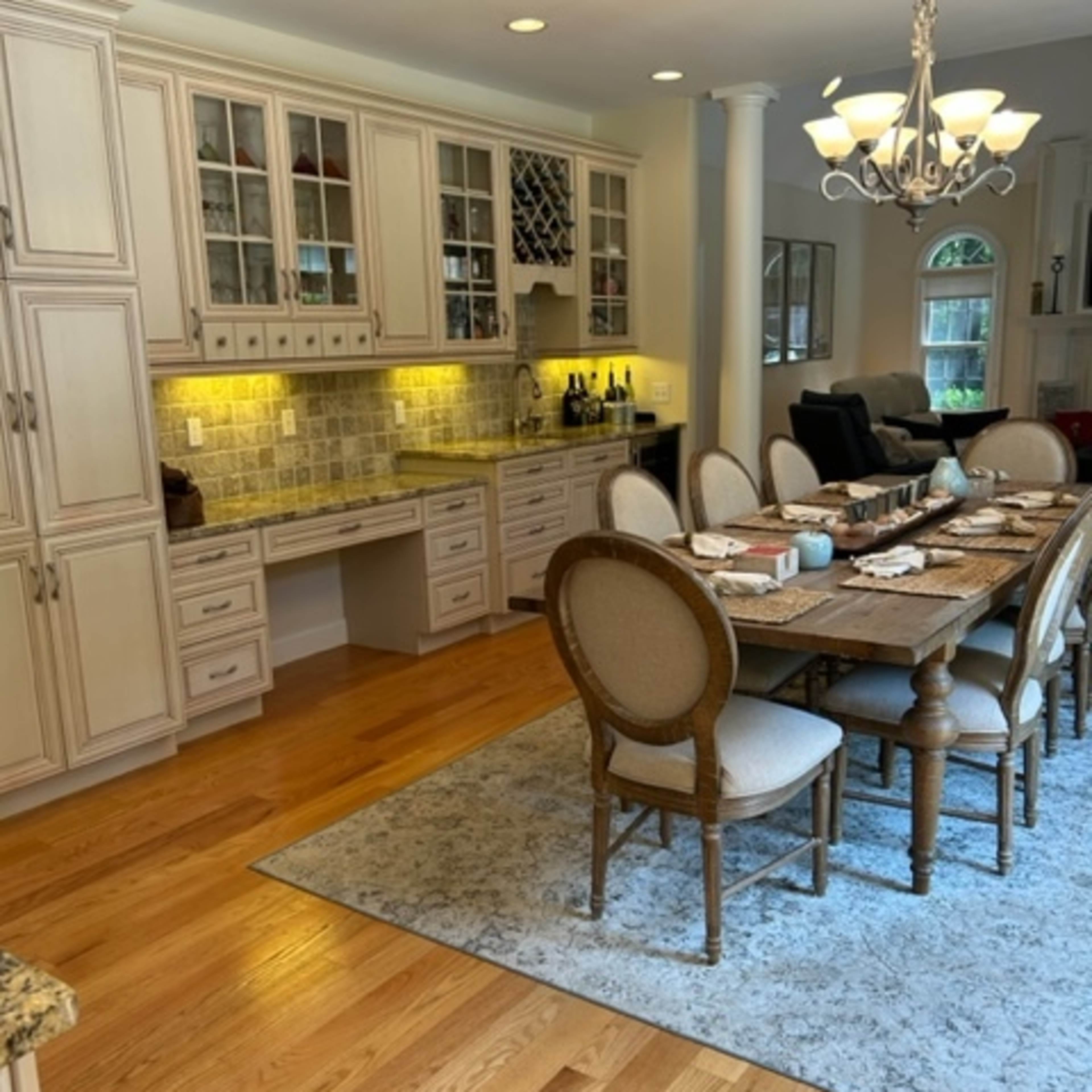 A spacious kitchen features a long dining table surrounded by upholstered chairs, with cabinets above and a countertop illuminated by under-cabinet lighting.