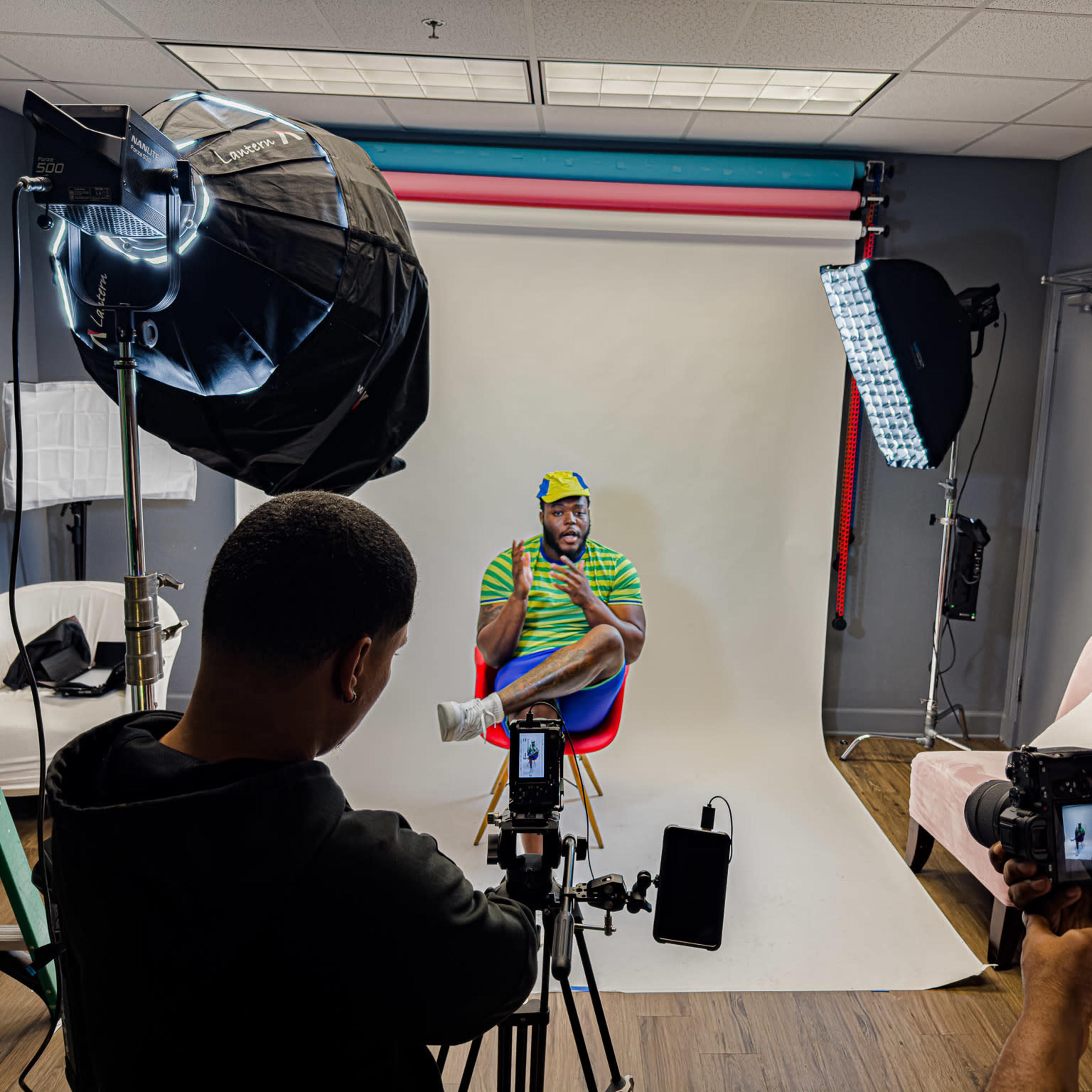 A man sits on a stool in front of a backdrop while being photographed by two individuals holding cameras in a well-lit studio.