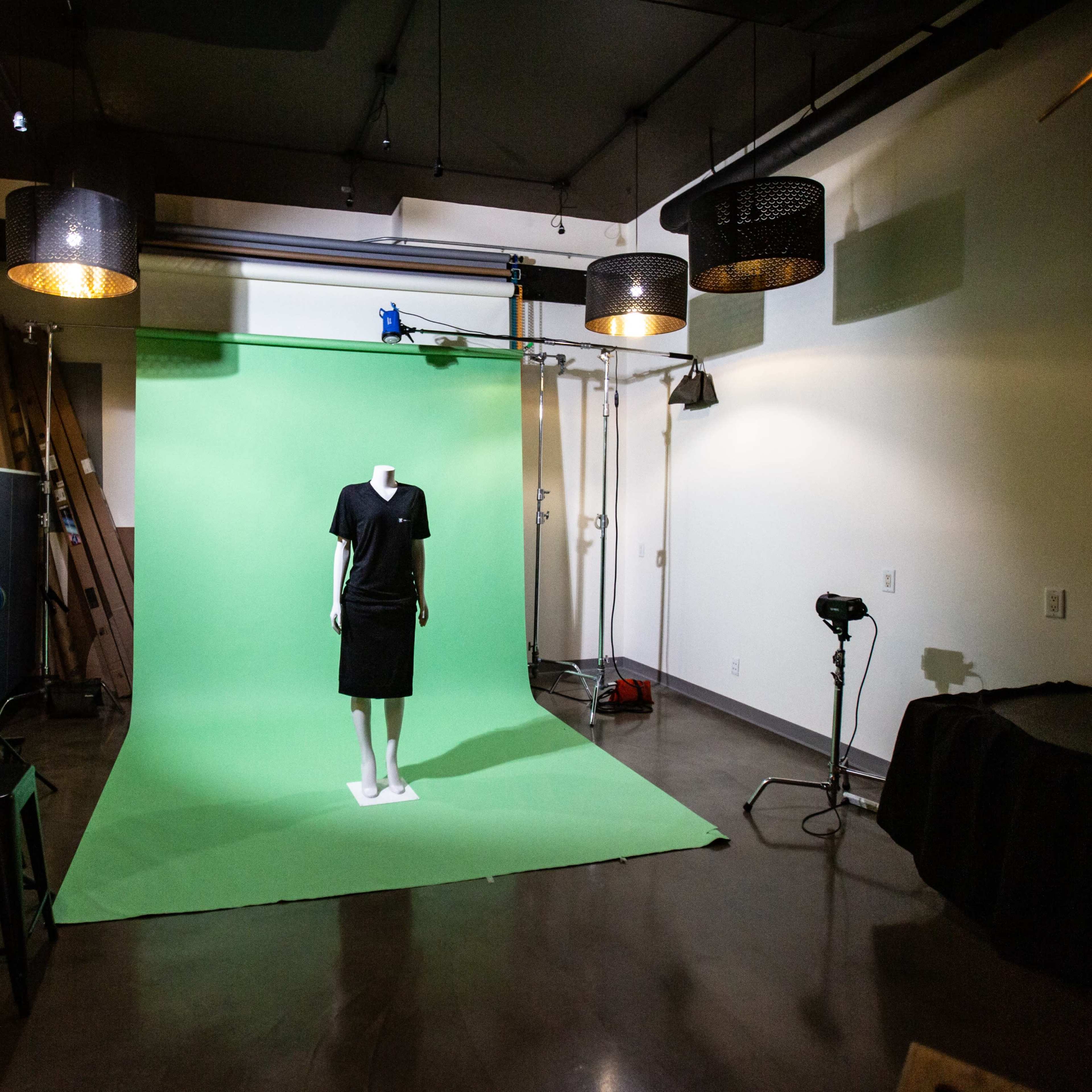 A mannequin dressed in a black outfit stands in front of a green backdrop, surrounded by studio lights and equipment in a photo studio.
