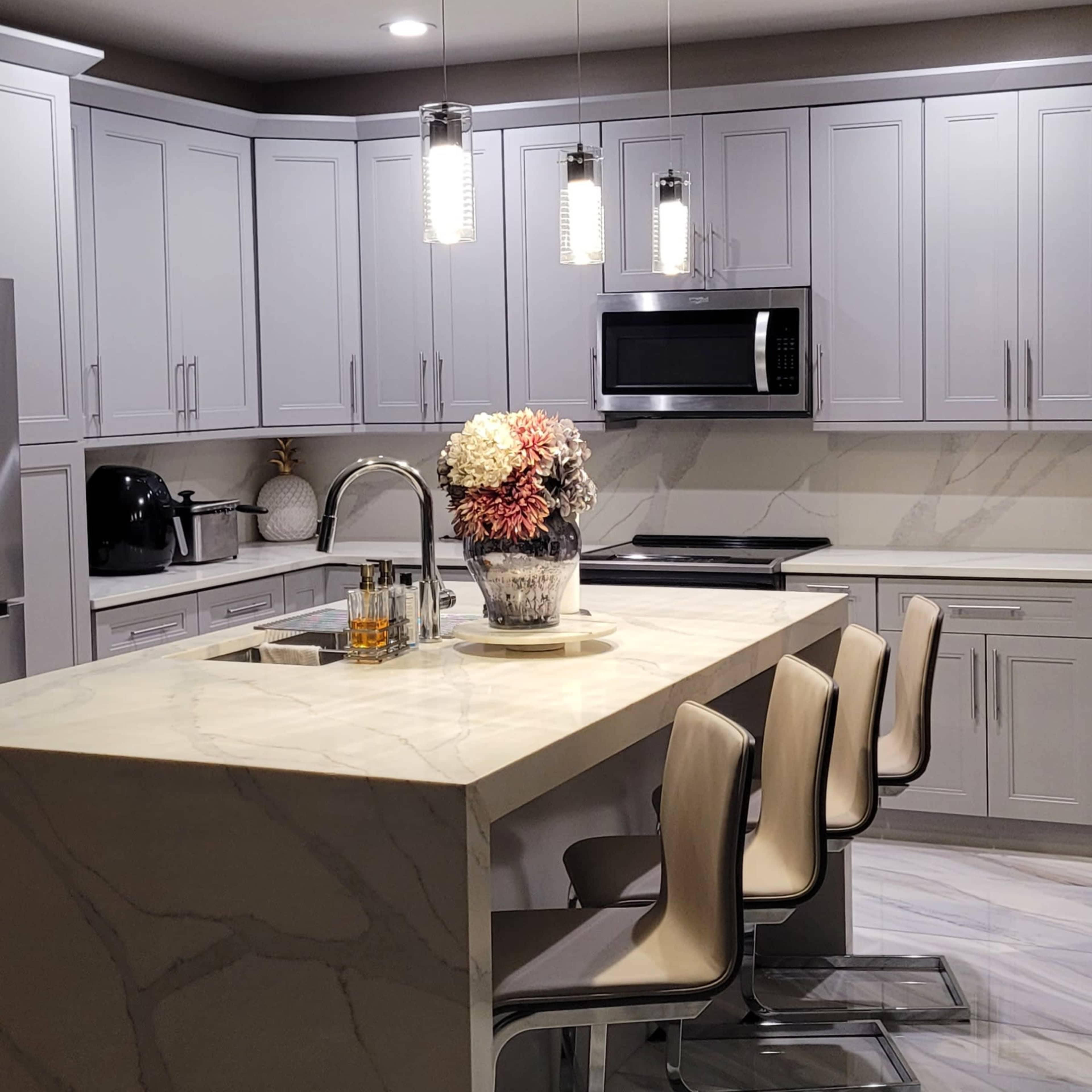 A modern kitchen with gray cabinetry, a marble island, and three bar stools, illuminated by pendant lights.