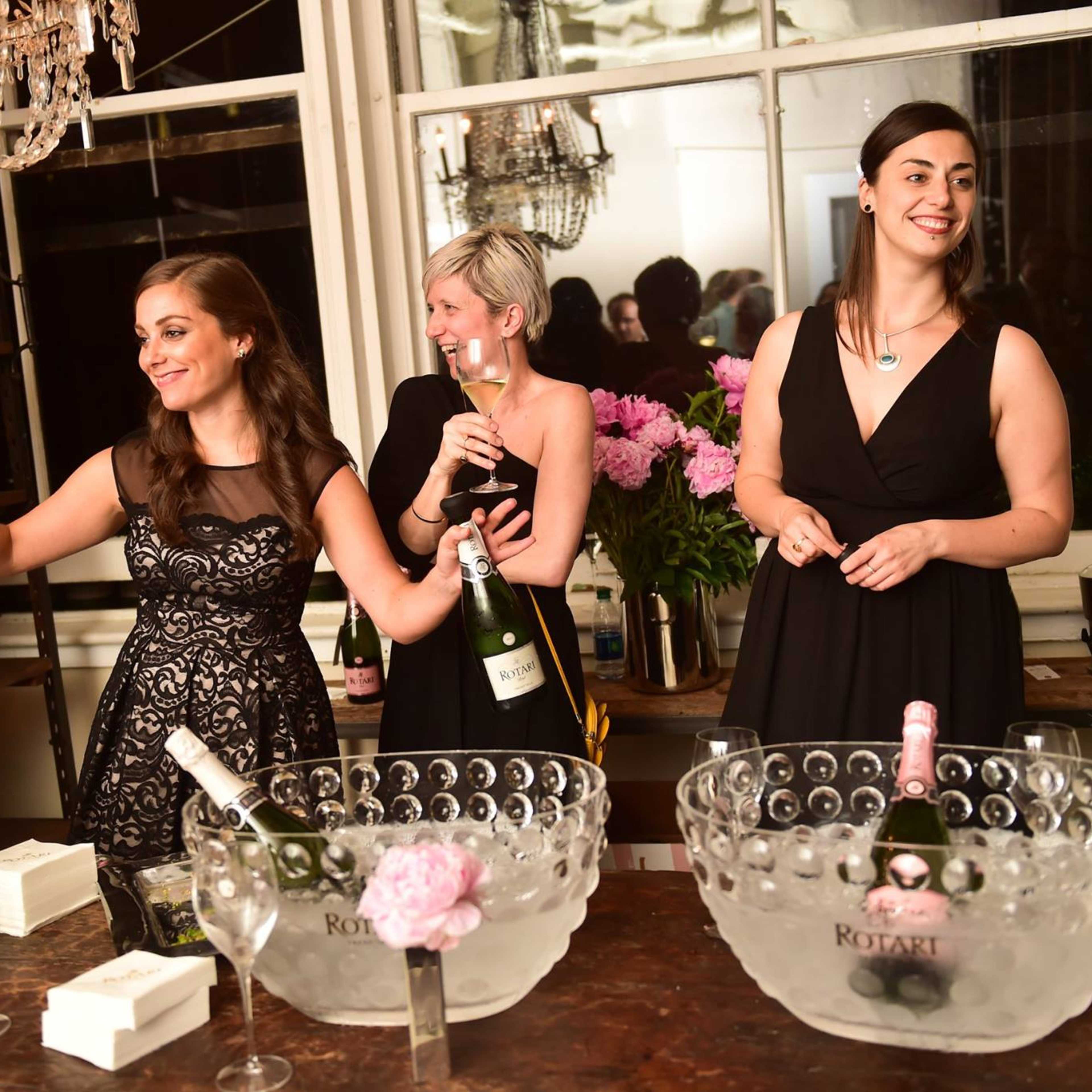 Three women in black dresses stand near two ice-filled bowls of champagne, with one woman gesturing and another holding a glass, in a room adorned with a chandelier.