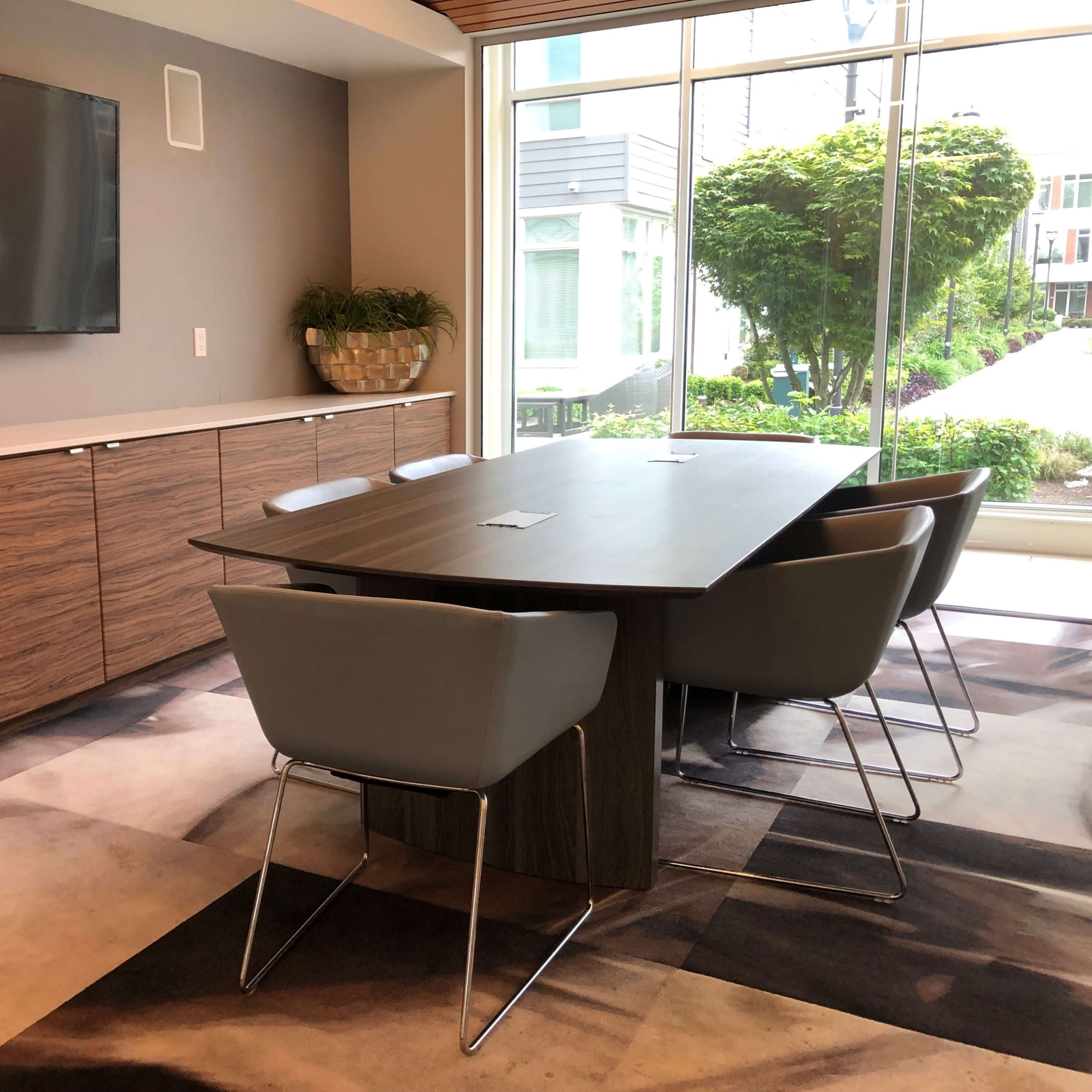 A modern conference room features a large wooden table surrounded by gray chairs, with a wall-mounted TV and large windows overlooking greenery.