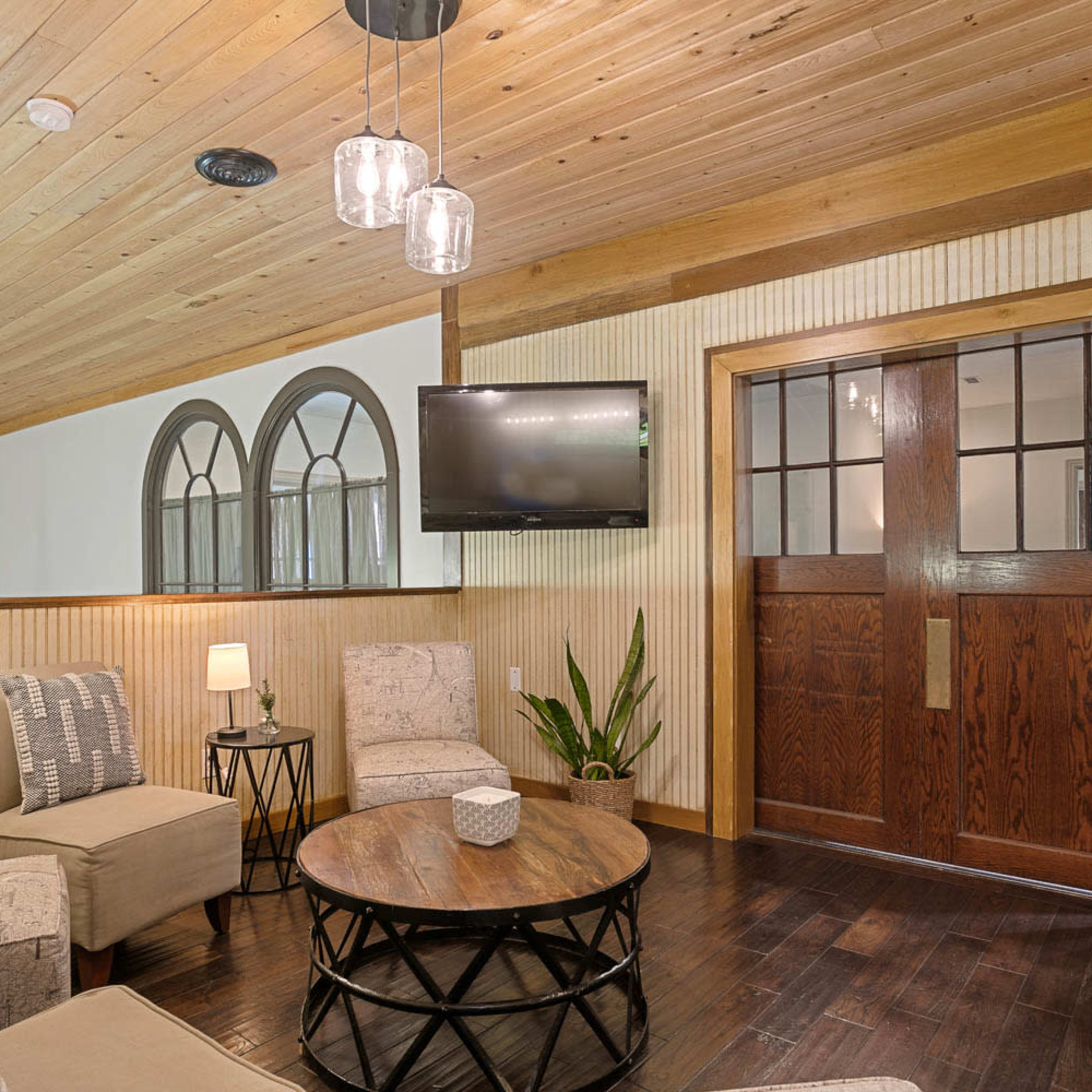 A living area with two armchairs, a circular coffee table, a flat-screen TV mounted on the wall, and a wooden front door with windows.