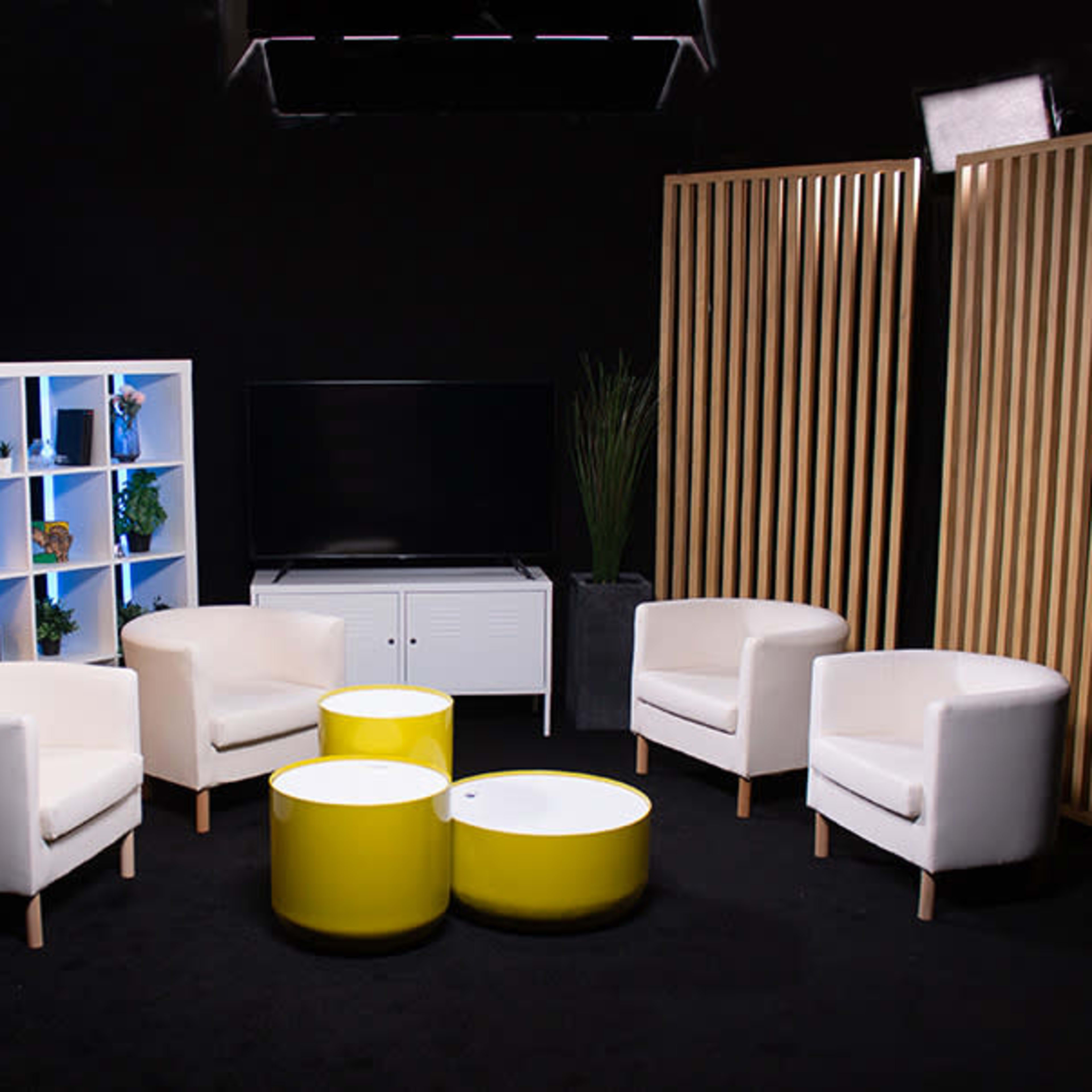 A modern studio setting with four white chairs arranged around two yellow coffee tables, a bookshelf, and a television on a low cabinet, all under soft lighting against a black backdrop.