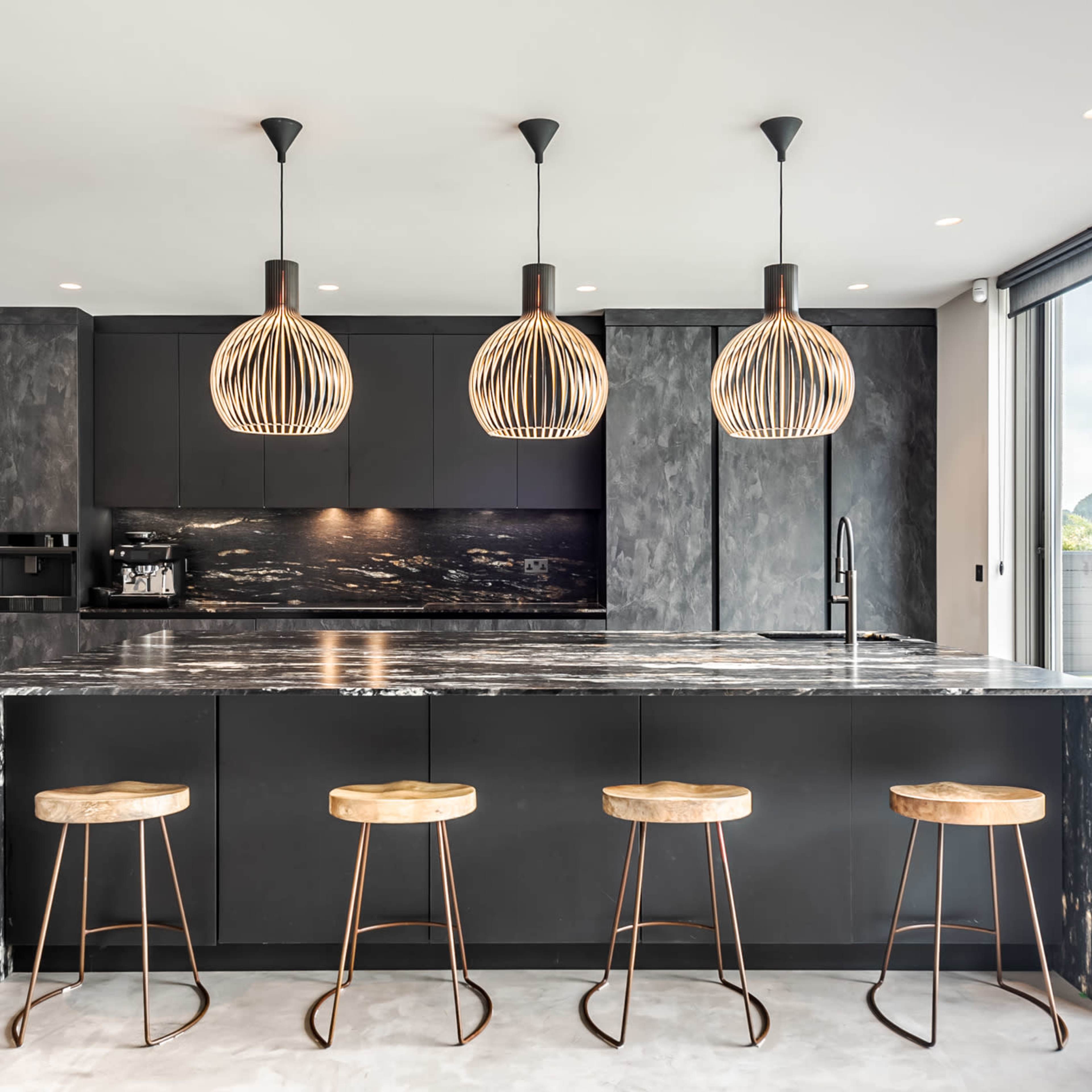 A modern kitchen featuring a dark cabinetry layout, a large island with a marble countertop, and three pendant lights above it, accompanied by four wooden stools.