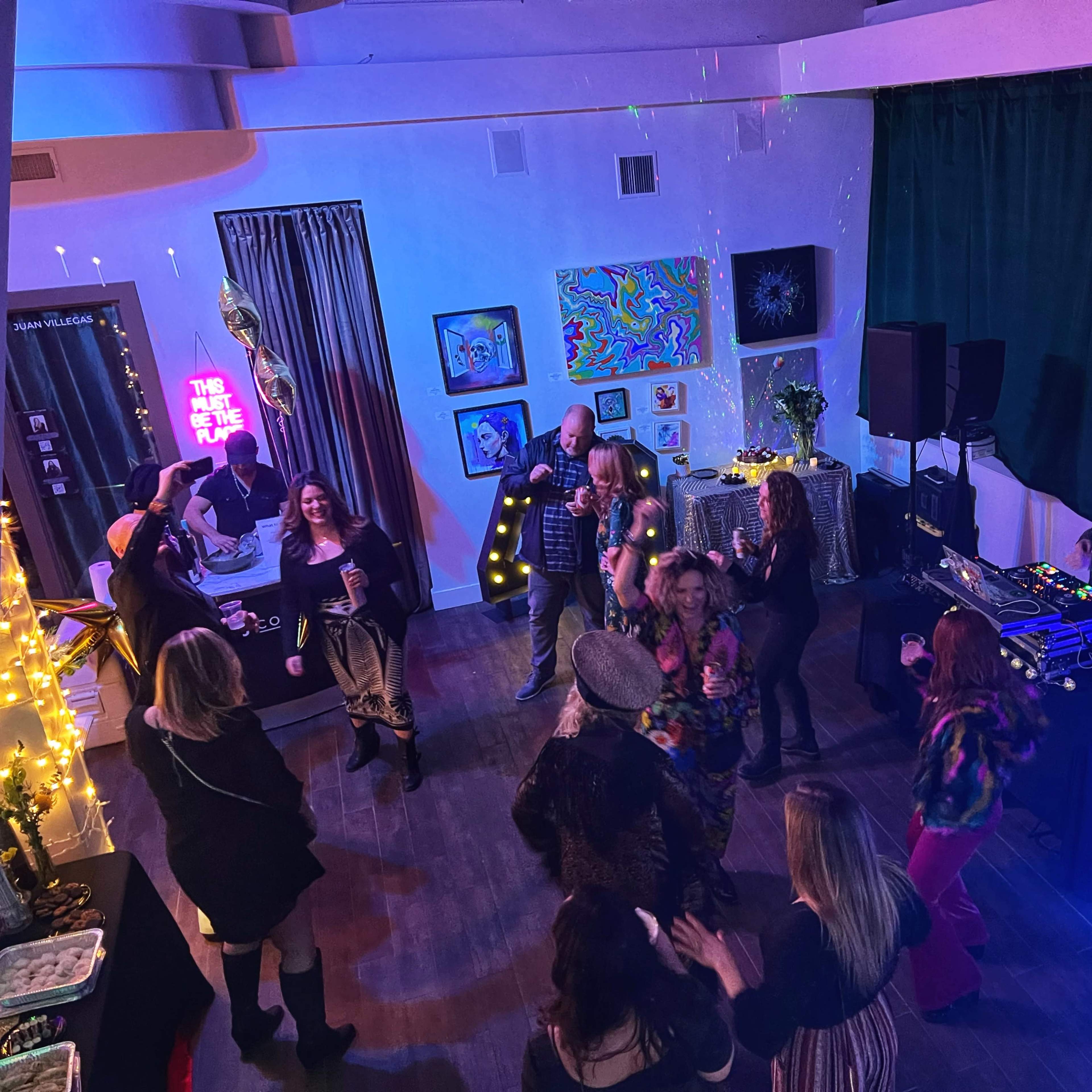 A lively group of people dance and socialize in a brightly decorated room featuring artwork and a DJ setup.